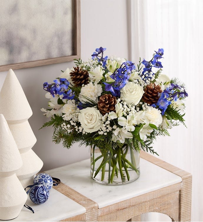 Blue Winter Whisper - An elegant winter bouquet of white roses, soft seasonal blooms, fresh evergreens, pinecones, and delicate blue accents in a clear glass vase.