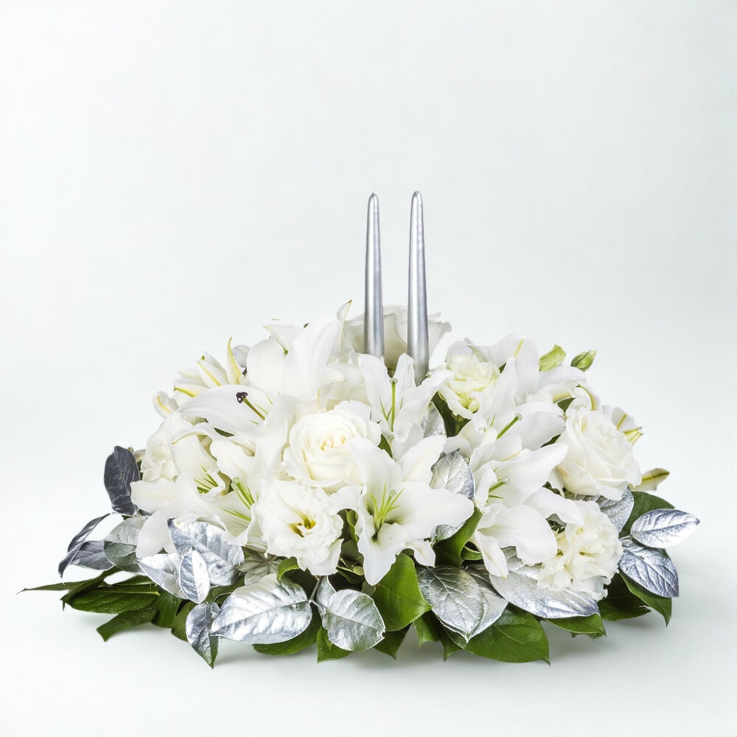 Winter Radiance Centerpiece - Elegant and serene, the Winter Radiance Centerpiece sets the tone for a refined holiday celebration. Featuring white lilies, roses, and lisianthus nestled among lush greenery and shimmering silver accents, this arrangement is crowned with two sleek silver taper candles. Perfect for holiday gatherings, seasonal décor, or a graceful gift, it brings a luminous touch to any table.  Picture Shows Deluxe Arrangement  What’s included: White lilies White roses White lisianthus Silver and green foliage Two silver taper candles Low centerpiece design