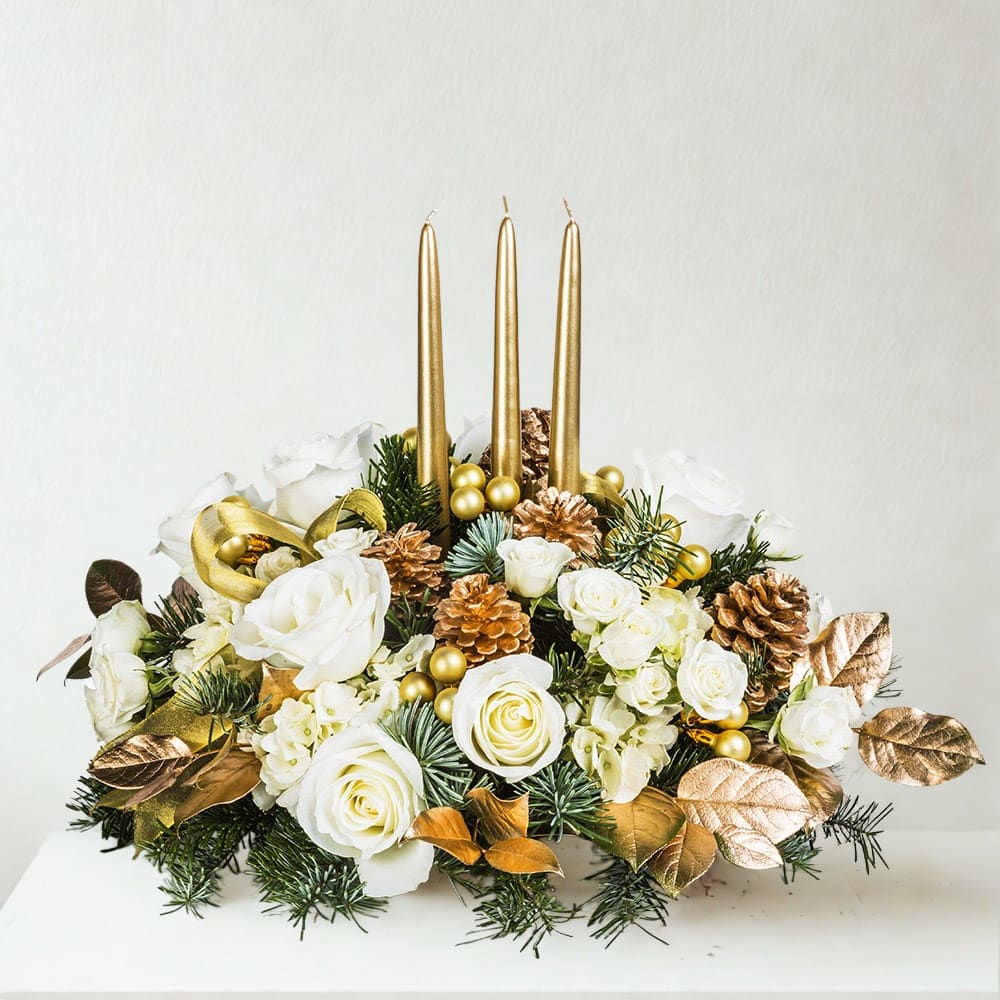 Golden Glow Centerpiece - Set an elegant tone with the Golden Glow Centerpiece—an opulent arrangement featuring lush white roses, creamy spray roses, snow-white hydrangeas, and festive greenery. Metallic gold and copper accents, including pinecones, leaves, and ornaments, bring warmth and radiance to the display. Three tall gold taper candles rise from the center, making this a sophisticated focal point for any table or gathering. Picture shows Deluxe Arrangement  What’s Included: White roses, spray roses, white hydrangea, evergreens, gold ornaments, gold pinecones, metallic foliage, gold taper candles