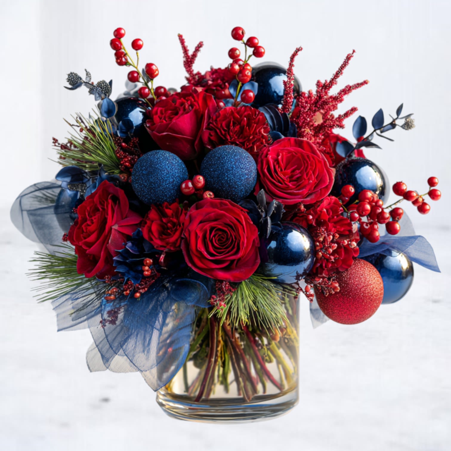 Grand Midnight Merriment - Turn up the festive drama with Grand Midnight Merriment, the elevated, fuller, and more decadent version of our bestselling Midnight Merriment design. Overflowing with premium red roses, red carnations, lush seasonal greenery, bold holiday berries, and an abundance of midnight-blue and crimson ornament spheres, this arrangement delivers show-stopping color and dimensional sparkle from every angle.  Designed in a modern clear glass vase with layered blue ribbon accents, this statement piece blends moody elegance with playful holiday shine. Perfect for entryway tables, holiday hosting, office gifting, or anyone who loves a vibrant, non-traditional Christmas palette.  Handcrafted in-house with premium blooms and layered textures for maximum fullness, height, and visual impact.  Available for same-day delivery in Magnolia, The Woodlands, Montgomery, Conroe, Tomball, and surrounding Houston areas.