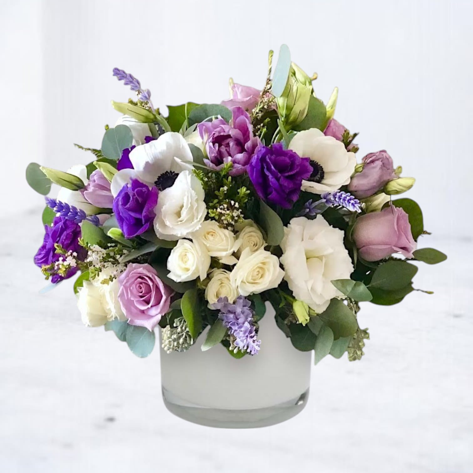 Periwave - Periwave is a contemporary floral design that blends soft movement with deep cool tones. Designed in a frosted white vessel, this arrangement features lavender roses, white roses, purple lisianthus, white anemones with dark centers, waxflower, eucalyptus, and sprigs of lavender for an added touch of aromatic calm.  Ideal for modern sympathy arrangements, or contemporary home décor, this design brings quiet sophistication to any space.