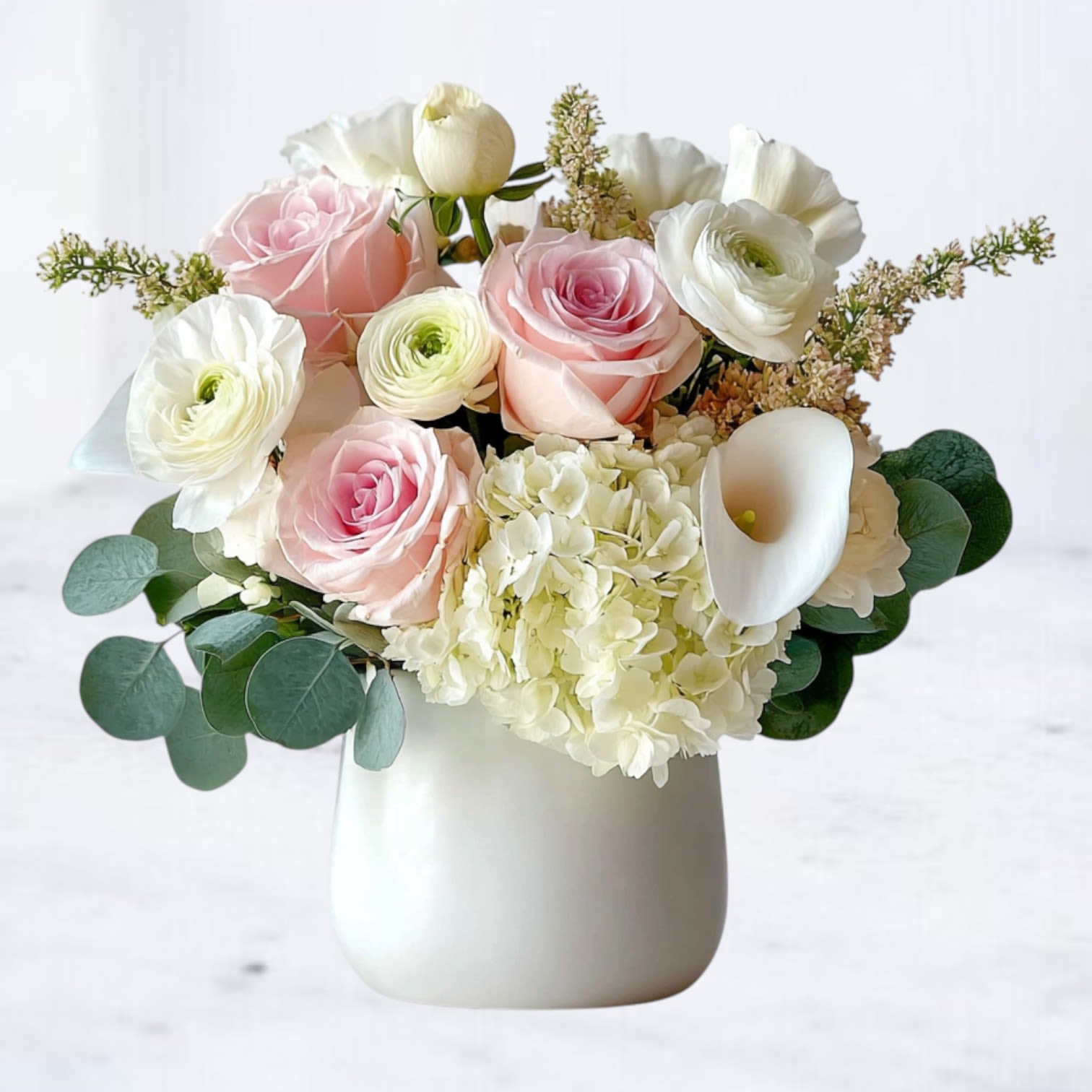Blush Whisper Bouquet - A delicate harmony of soft blush and creamy white blooms, the Blush Whisper Bouquet captures timeless elegance in every petal. Featuring pale pink roses, white ranunculus, lush hydrangea, and graceful calla lilies, this arrangement is designed in a modern white ceramic vase with fresh eucalyptus for texture and balance. Perfect for birthdays, anniversaries, sympathy gestures, or simply brightening someone’s day, this bouquet brings quiet sophistication to any space. Available for same-day delivery in Magnolia, The Woodlands, Spring, Montgomery, and surrounding areas.