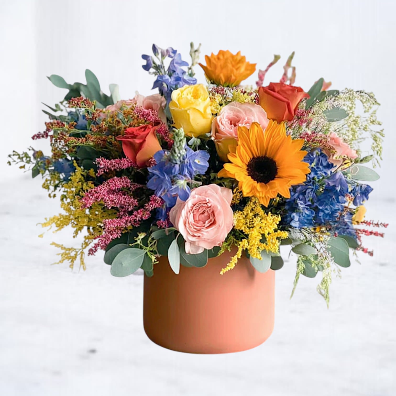 Cheerful Drop - Capture the essence of a sunlit garden with Cheerful Drop, a vibrant floral arrangement that radiates warmth and joy. Designed in a chic matte terracotta vase, this breathtaking bouquet features golden sunflowers, peach garden roses, deep blue delphinium, and delicate yellow alstroemeria, accented with lush greenery and textural elements for a naturally elegant touch.  Perfect for birthdays, celebrations, or simply brightening someone's day, Cheerful Drop brings a lively and organic beauty to any space. Available for same-day delivery in Houston and surrounding areas, this arrangement is a stunning way to share happiness and love.