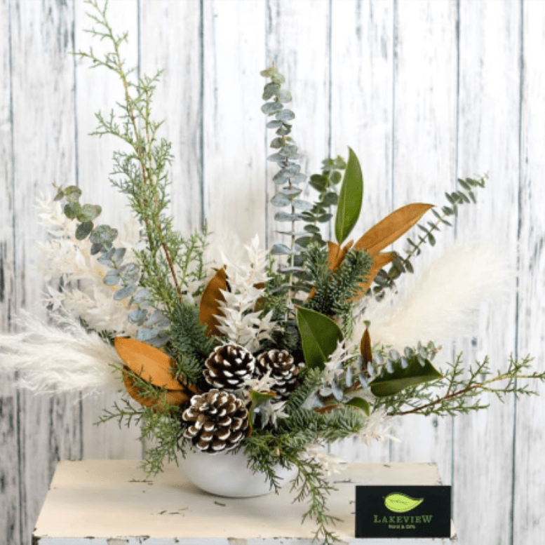 Urban - Embody the quiet magic of a winter forest with this elegant, nature-inspired arrangement. Designed in a modern white bowl, this piece blends soft neutrals with rich greenery for a look that feels both fresh and timeless. Frost-kissed pinecones sit at the heart of the design, surrounded by textured elements such as eucalyptus, feathery pampas plumes, cedar, and evergreens. Accents of magnolia leaves add warmth and contrast, creating a beautifully layered composition.  Perfect for holiday tables, entryways, or seasonal gifting, this arrangement evokes the calm, cozy charm of winter while maintaining a clean, contemporary aesthetic. Handcrafted with care by Lakeview Floral &amp; Gifts, it’s a natural statement piece that celebrates the season with understated elegance.  Approximate Dimensions: 15&quot;W x 15&quot;T