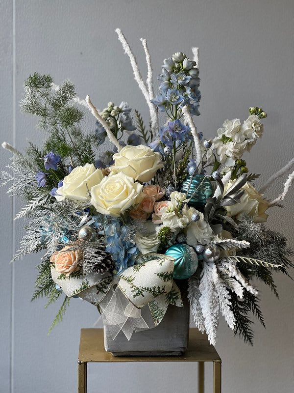 Antarctic Blizzard - Our &quot;Antarctic Blizzard&quot; Arrangement is a frosty cool tone arrangement that feature's; Snow branches, blue delphinium, white roses, white stock, and snow greenery finished in a cement vase. This design connotates towards the feeling of happiness one feels while riding on a sled and the memories of this season's winter.