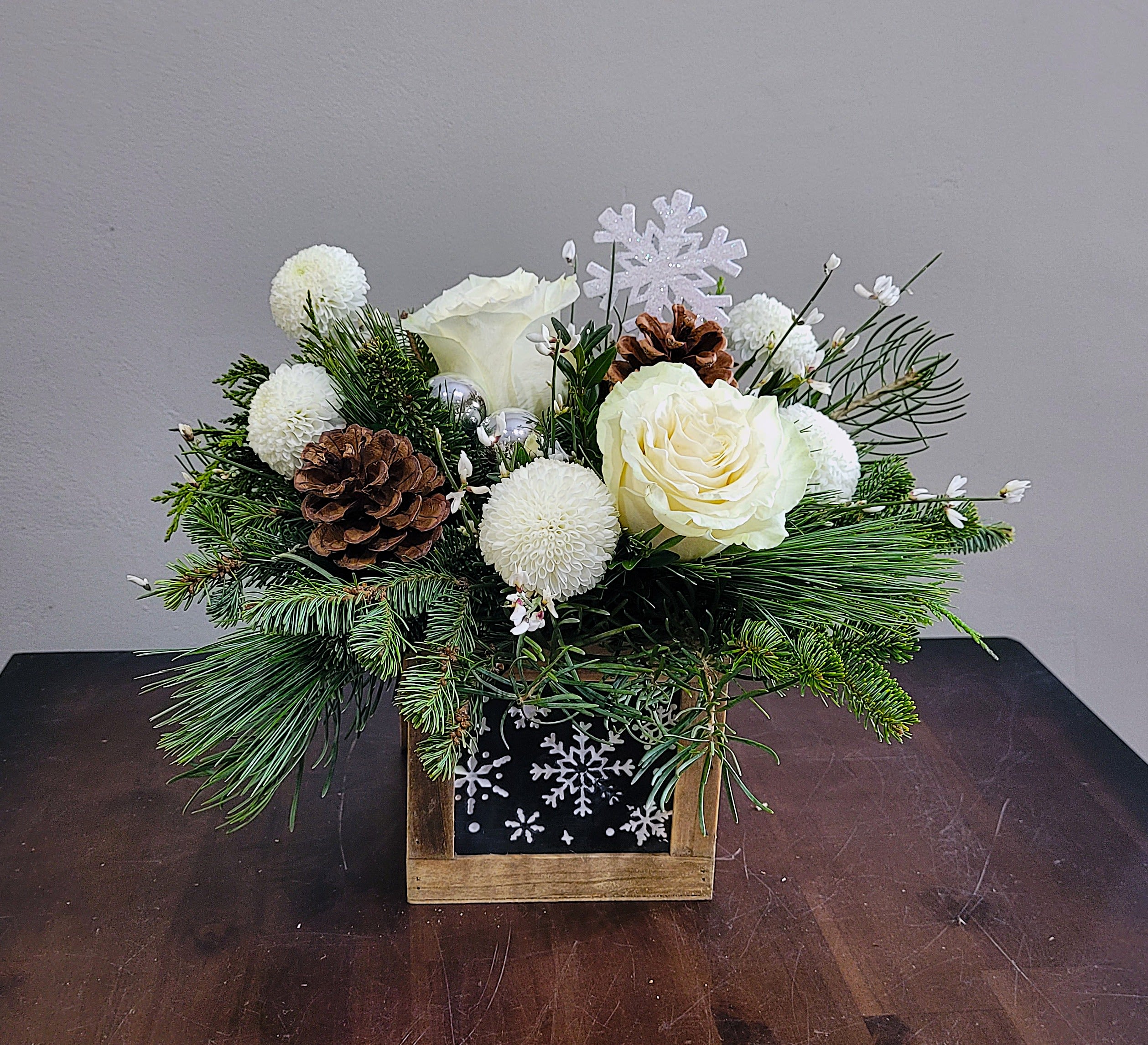 Snowdrop Bouquet - Hammered tin snowflakes are the winters charm in a rustic wood box. A mix of evergreen pines surrounds white roses, button mums and genista accented with festive baubles and pinecones. 