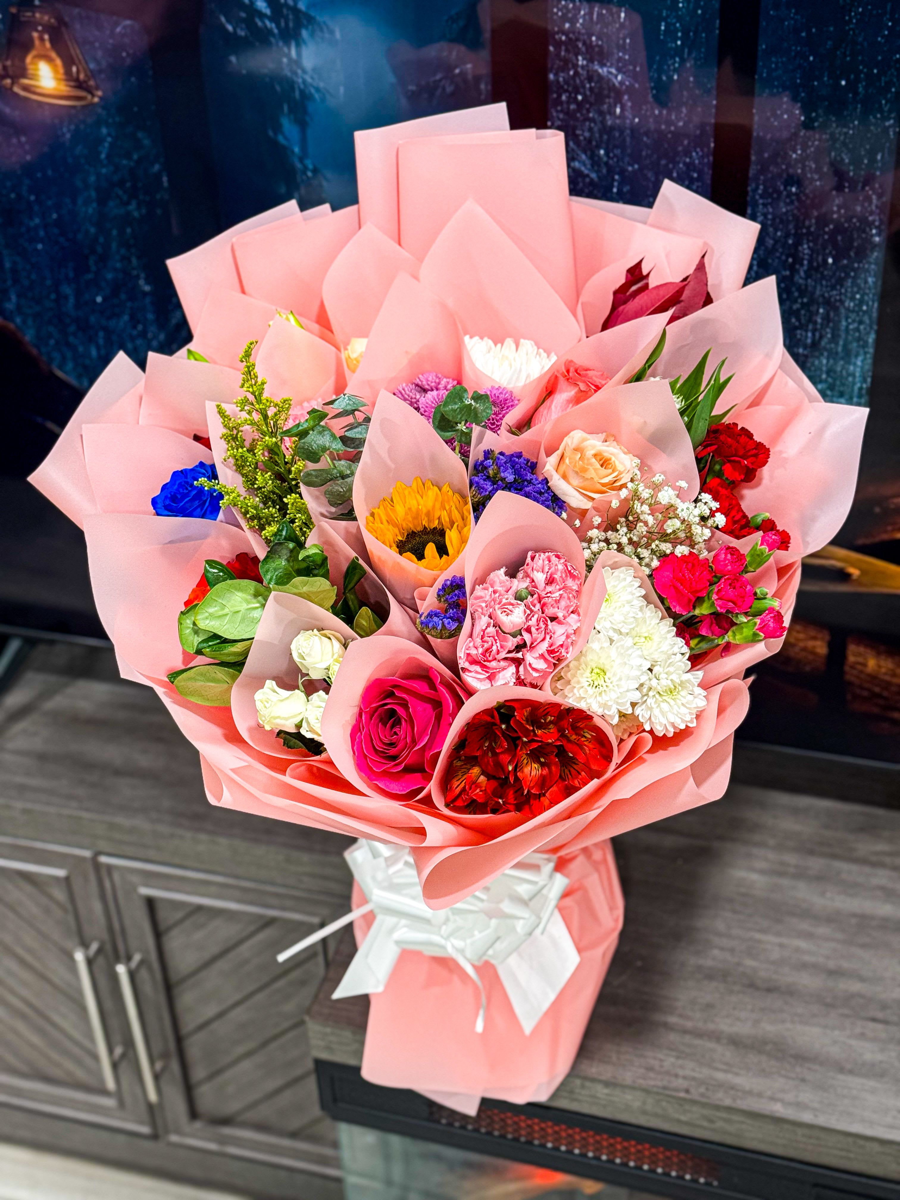 Blush Garden Grande - is a stunning hand-crafted bouquet wrapped in our signature Korean-style blush wrap. Designed with a mix of premium seasonal blooms, this arrangement brings together vibrant roses, sunflowers, chrysanthemums, carnations, and lush greenery — each flower placed with purpose to create depth, color balance, and elegance.  Perfect for birthdays, anniversaries, congratulations, or simply making someone feel special. This bouquet is made to impress — big, full, and picture-perfect from every angle.