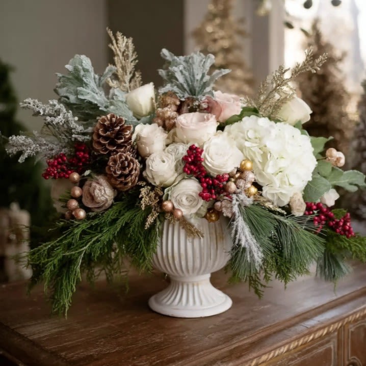 Christmas Elegance - An elegant christmas floral arrangement in a classic urn, with winter roses, berries, and evergreens glowing softly by candlelight.  There’s a certain magic that happens when old-world charm meets the hush of winter. This pedestal urn is our ode to that feeling: a graceful vessel overflowing with creamy garden roses, plush hydrangea, snow-dusted pine, silvery eucalyptus, and cinnamon-kissed pinecones. Ruby berries play against the soft whites for a look that reads festive, not flashy. If your holiday style leans toward thoughtful textures and quiet glow, this piece will anchor your space with ease. It’s equally at home on a dining sideboard, entry console, or as the hero of your Christmas table centerpiece where conversation flows and candles sparkle.
