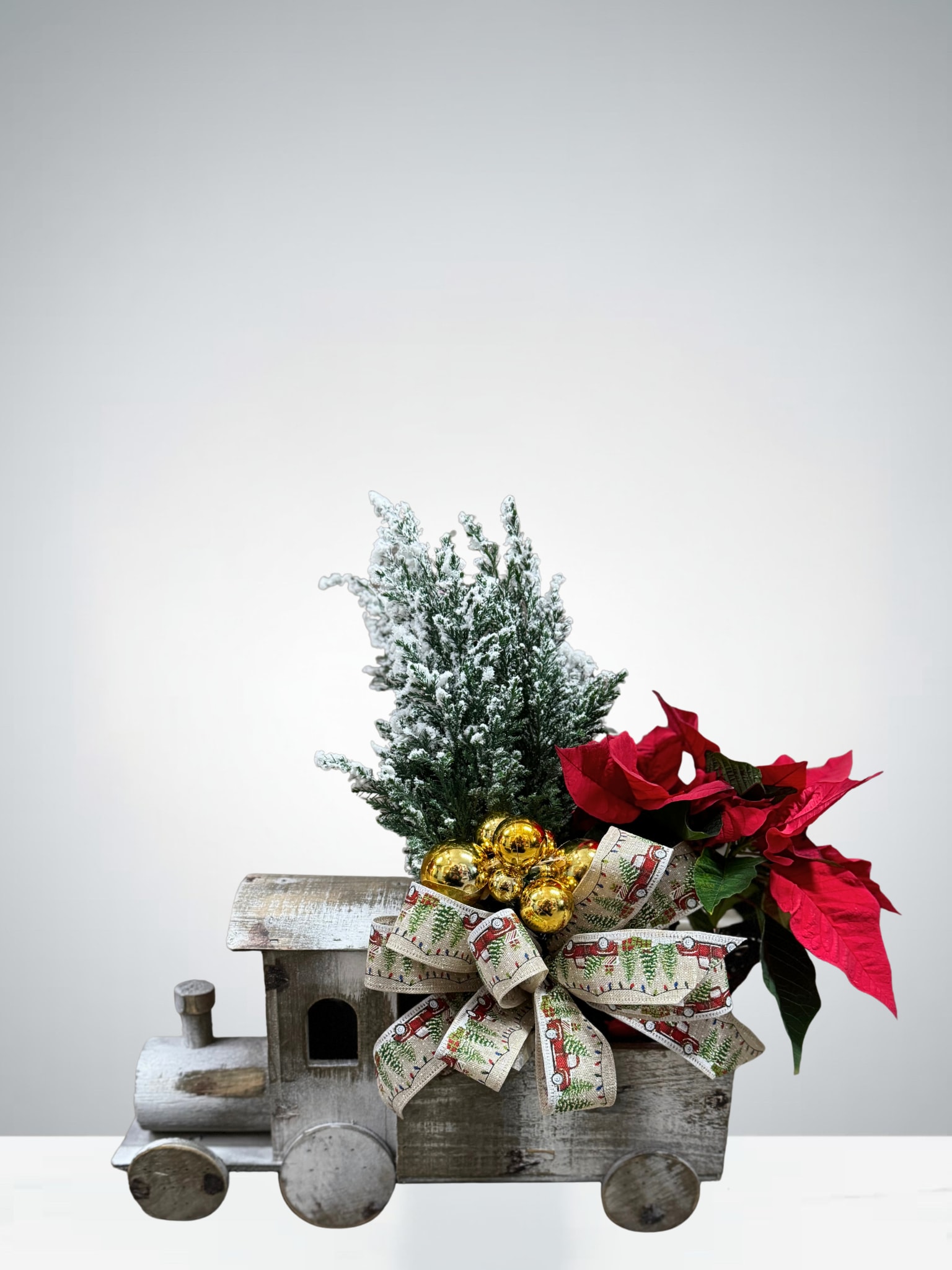 Christmas Express  - All aboard the holiday cheer! This charming arrangement features a small poinsettia paired with a tiny, snow-kissed cedar tree, all nestled inside a rustic wooden train planter. Adorned with festive ribbon and golden ornaments, it brings a playful and cozy Christmas touch to any space. Perfect for tabletops, entryways, or gifting a little extra holiday magic.