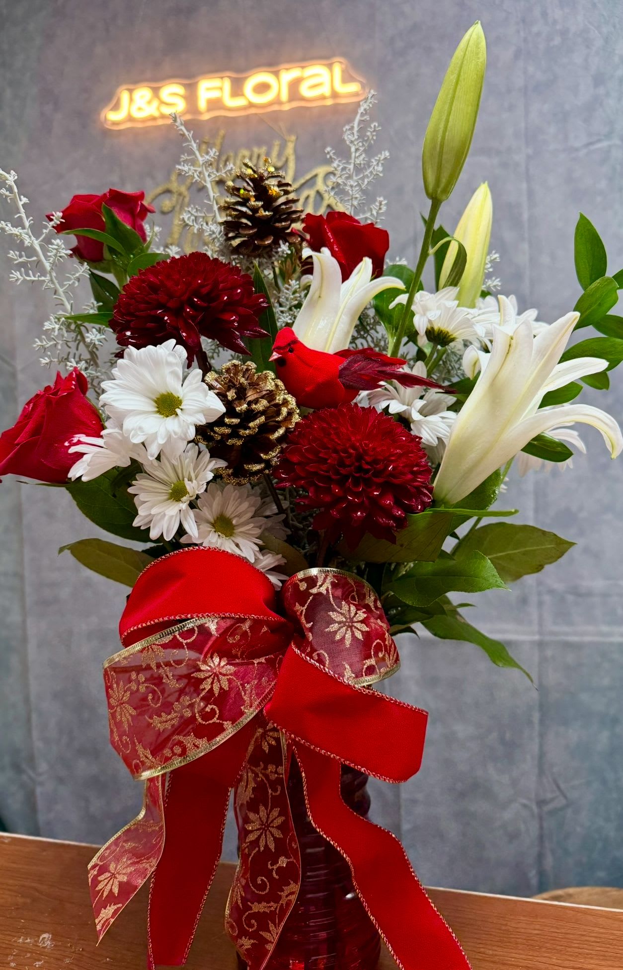 Cardinal Christmas Cheer - A joyful holiday vase featuring rich red blooms, snowy whites, fragrant lilies, and a charming red cardinal perched among gold-tipped pinecones.