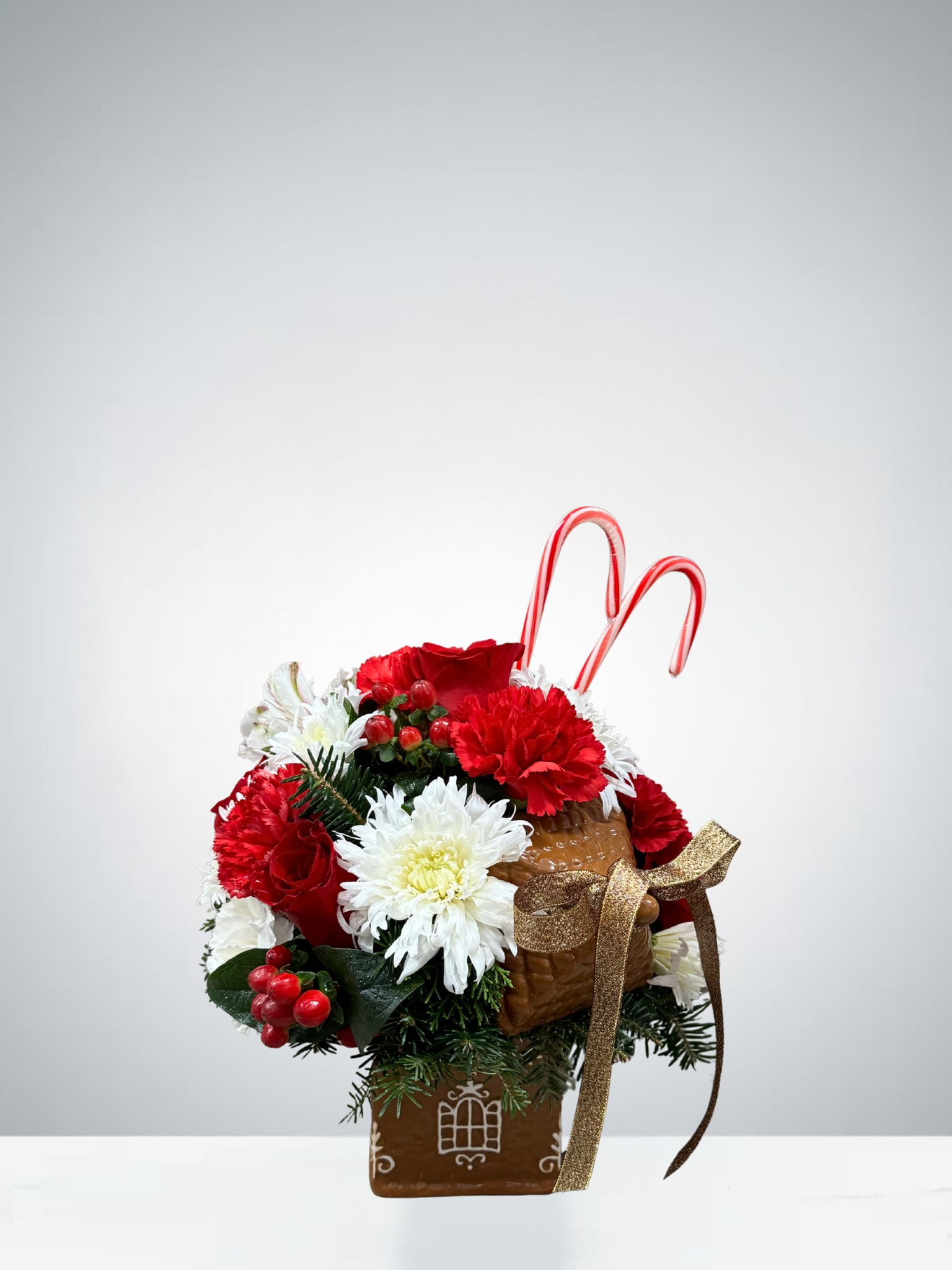 Gingerbread Cottage Arrangement - Bring home a touch of sweet holiday magic with this festive arrangement displayed in an adorable gingerbread-themed container. Filled with a joyful mix of red and white blooms, seasonal greenery, bright berries, and two cheerful candy canes, this piece captures the cozy charm of Christmas.
