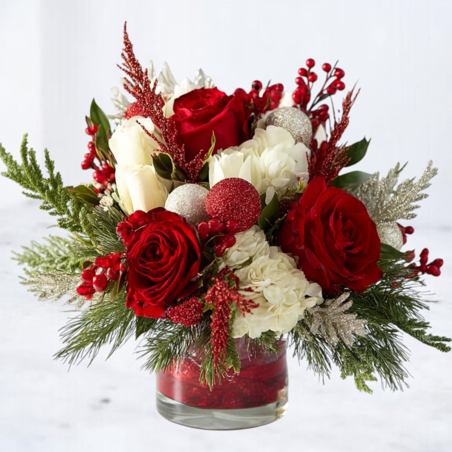 Yuletide Rouge - Bold, festive, and unmistakably luxurious, Yuletide Rouge is a show-stopping holiday arrangement designed to bring the magic of Christmas straight to your table. Deep red &amp; white roses, snowy white hydrangea, and winter-fresh evergreens come together with shimmering red and silver ornaments for a look that feels both classic and modern. Sparkling accents and pops of holly-red berries add texture and depth, while the glitter-lined vase completes the design with cheerful holiday sparkle.  Whether you’re hosting a Christmas dinner, elevating your holiday décor, or sending a heartfelt seasonal gift, Yuletide Rouge delivers the perfect blend of warmth, richness, and festive elegance.  Available for same-day Christmas flower delivery in Magnolia, The Woodlands, Conroe, Tomball, Spring, Montgomery, and surrounding Houston areas.