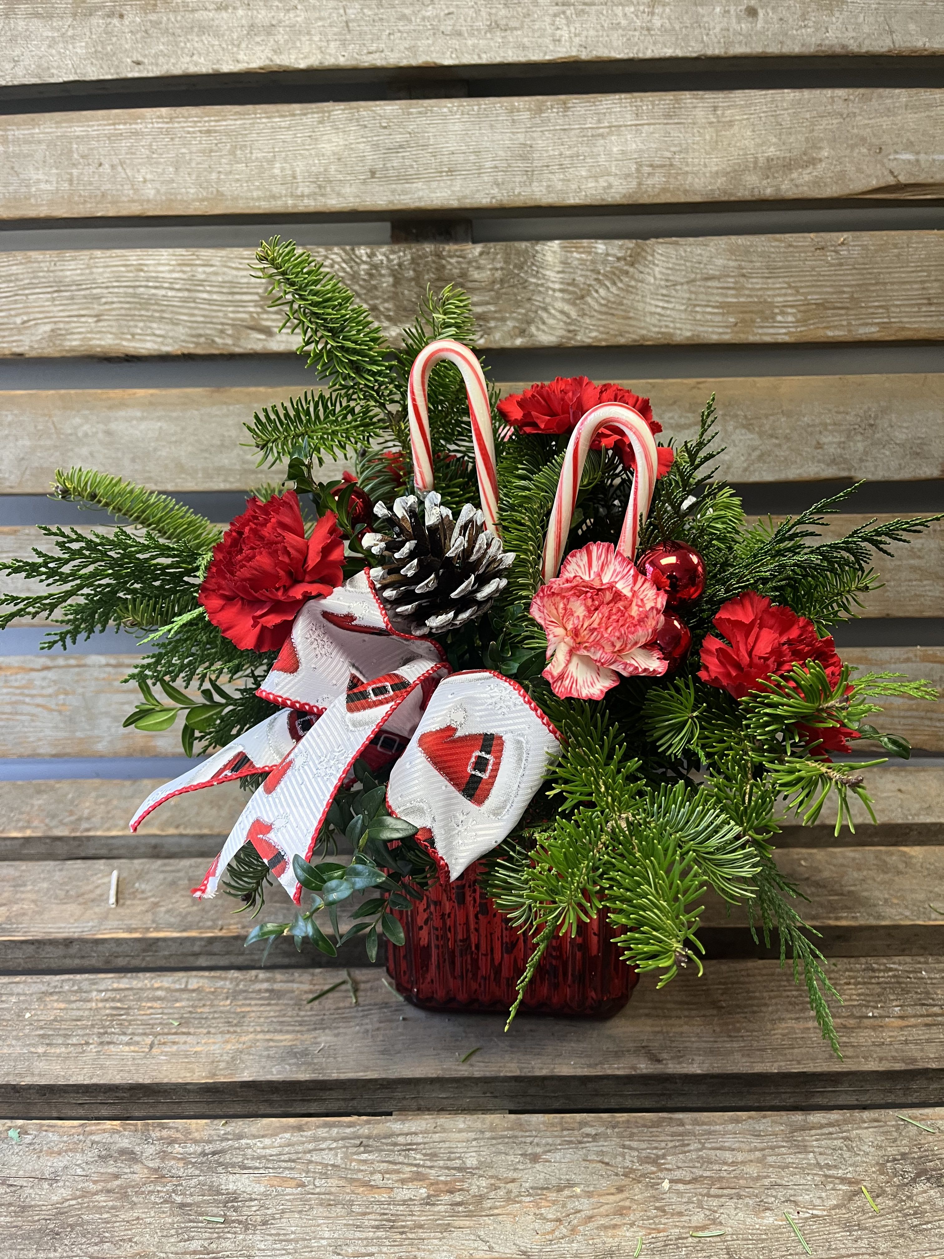 Candy cane lane  - Red Mercury cube filled with seasonal Christmas greens and pops of long lasting carnations. Accents of Santa ribbon, Christmas balls, candy, canes, and pinecones.