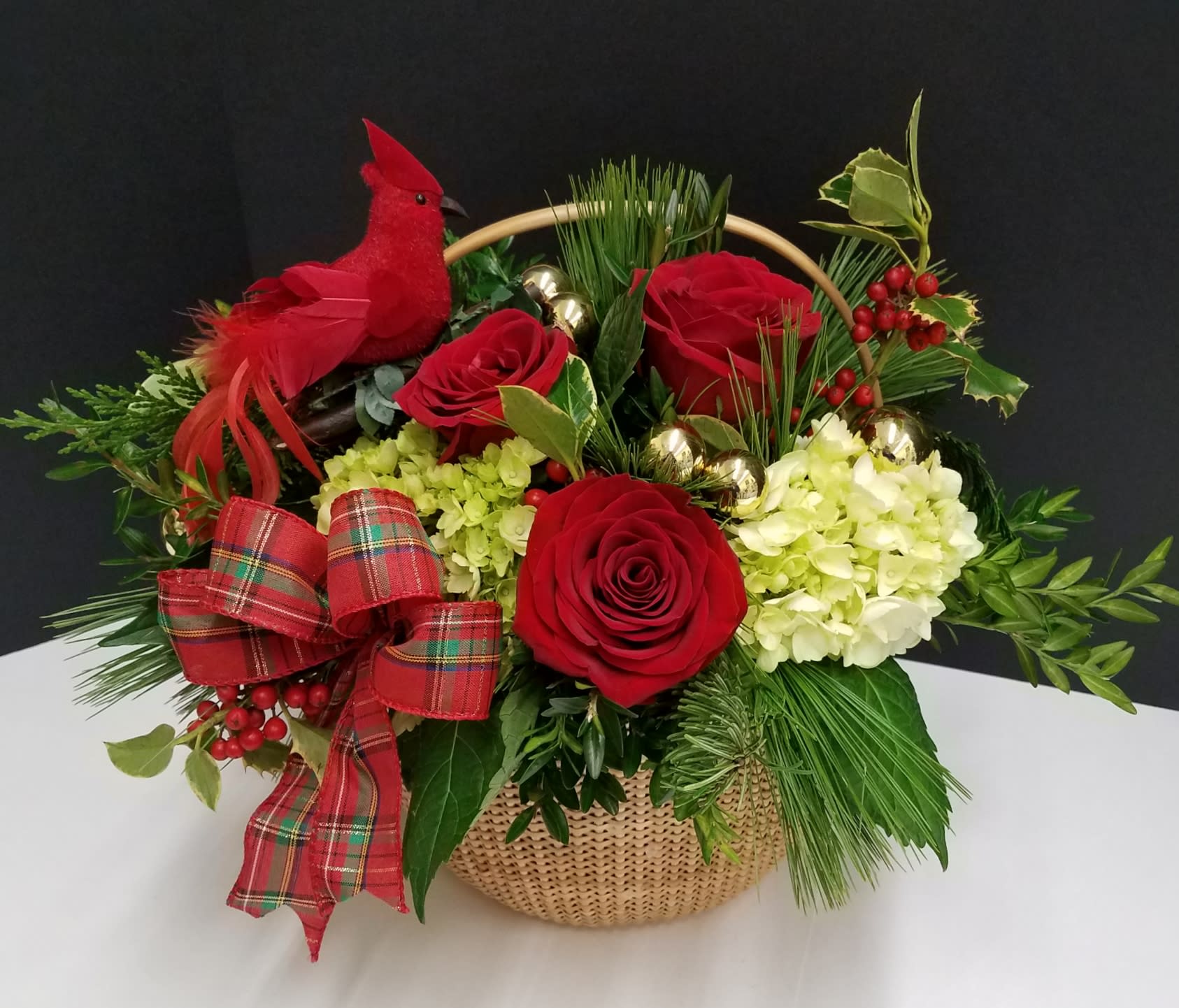 The Cardinal's Garden - A festive arrangement in a classic Nantucket basket adorned with a handsome cardinal perched in a garden of assorted winter pine, holly, hydrangea, red roses and finished with a beautiful plaid bow and gold ornament balls.