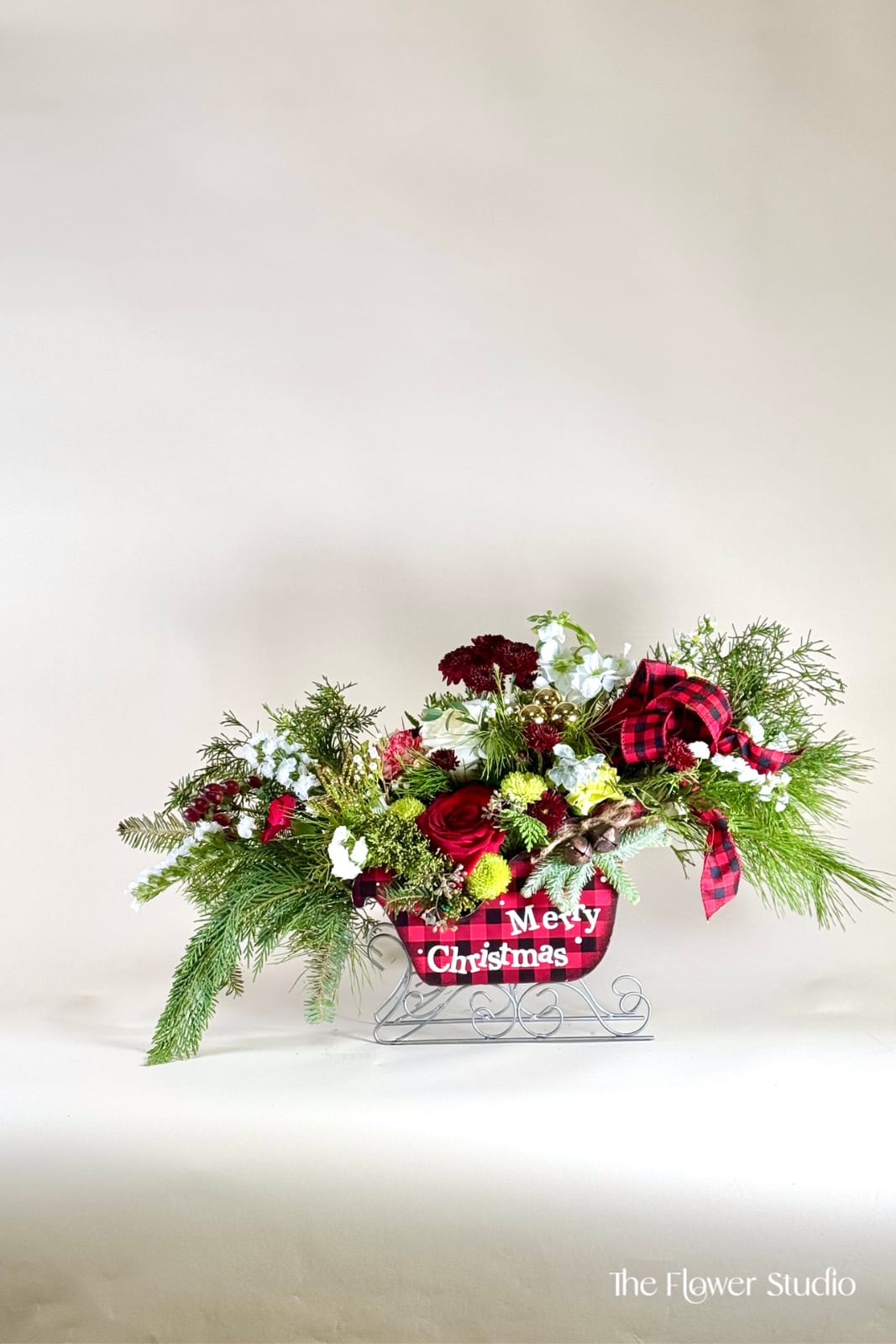 Sleighbell - Celebrate the season with this charming Christmas sleigh arrangement, overflowing with festive reds, snowy whites, winter greens, and cozy plaid accents. This sleigh it brings the magic of the holidays to any table or entryway. Lush textures, seasonal details, and a warm, rustic feel make this piece a perfect gift or holiday centerpiece. A cheerful and inviting design that captures the heart of Christmas.