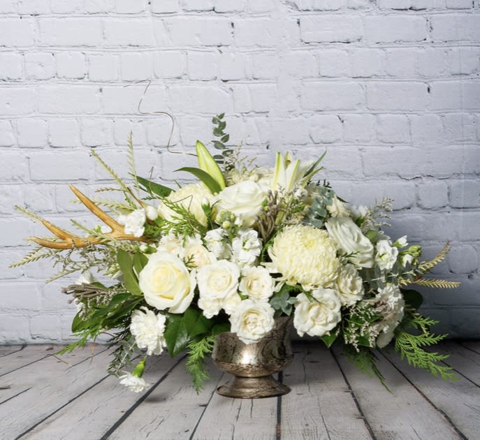 Winter Wonderland - An eye catching, gorgeous all white and gold arrangement in a antiqued gold compote. This stunning centerpiece with its antler accessory will impress! Comes with roses, spray roses, fragrant stock and lilies, chrysanthemums, silver brunia, pinecones, and wax flower. 