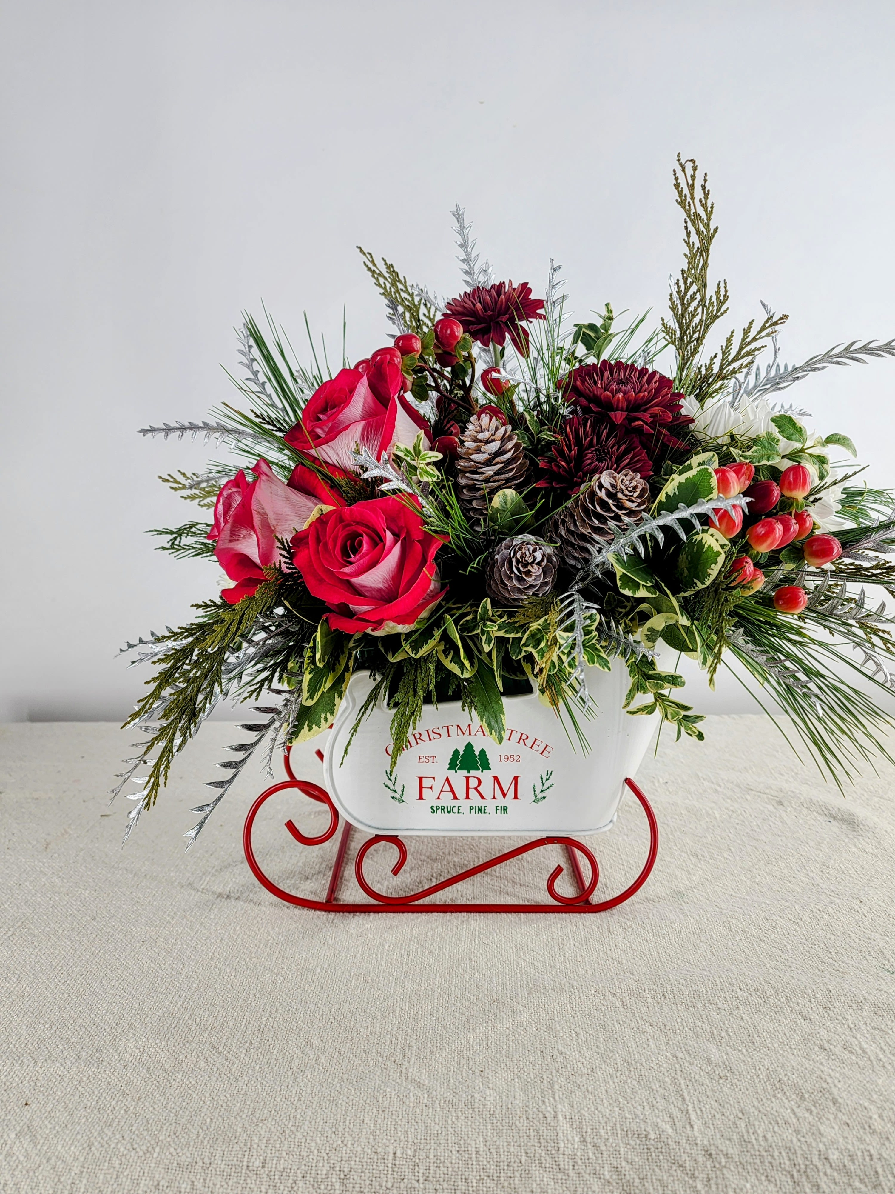 Sleigh Time - This festive sleigh embodies our Winter wonderland. It is adorned with roses, mums, hypericum berries, frosted pine cones, and a variety of evergreens. It serves as a delightful centerpiece to warmly greet family and friends during the holiday season.