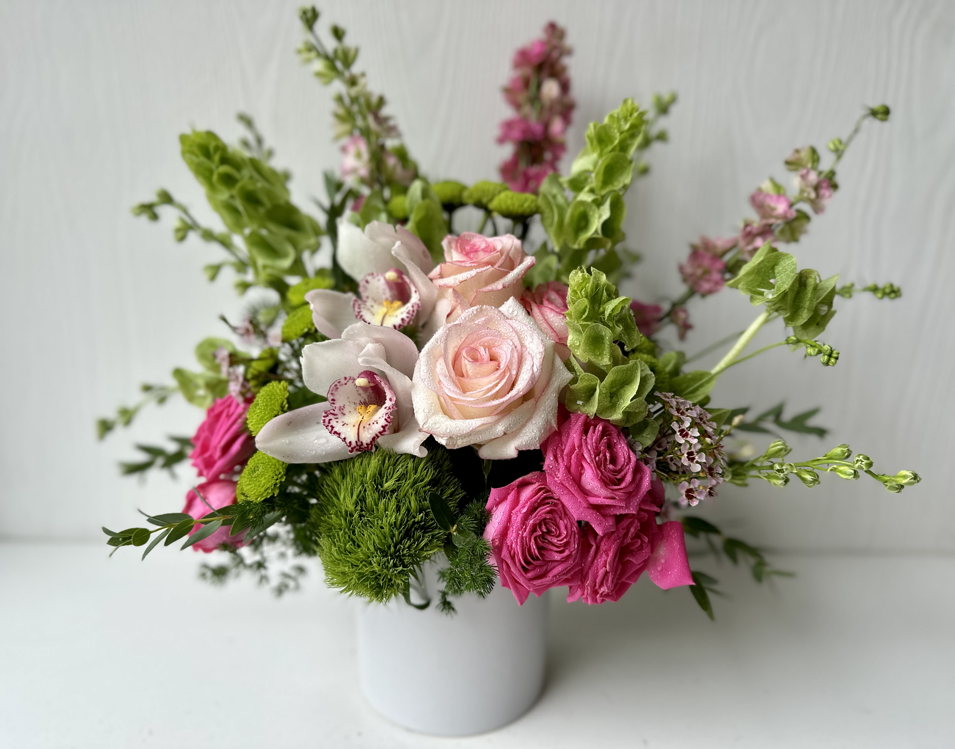 EC80 -Sweet in Pink - Escape to paradise with this lush and vibrant bouquet. This floral arrangement contains a design featuring fresh Cymbidium Orchids, Bells of Ireland, Snapdragon, Roses, and Button Mums. This is the perfect gift for a birthday, congratulations, or to celebrate new beginnings.   APPROXIMATE DIMENSIONS:  8" H x  7"W