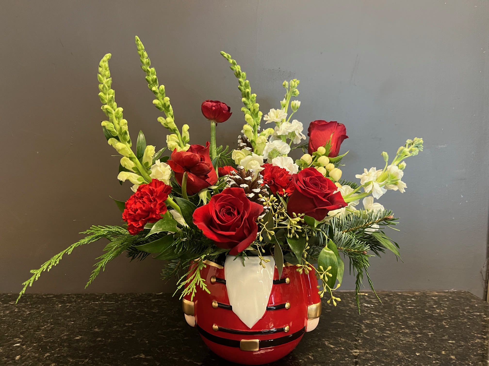 Mr. Nutcracker - this bouquet is a festive mix of Christmas greens, roses, ranunculus, snapdragons, stock, winter berries and pinecones in a cute little ceramic nutcracker container.  