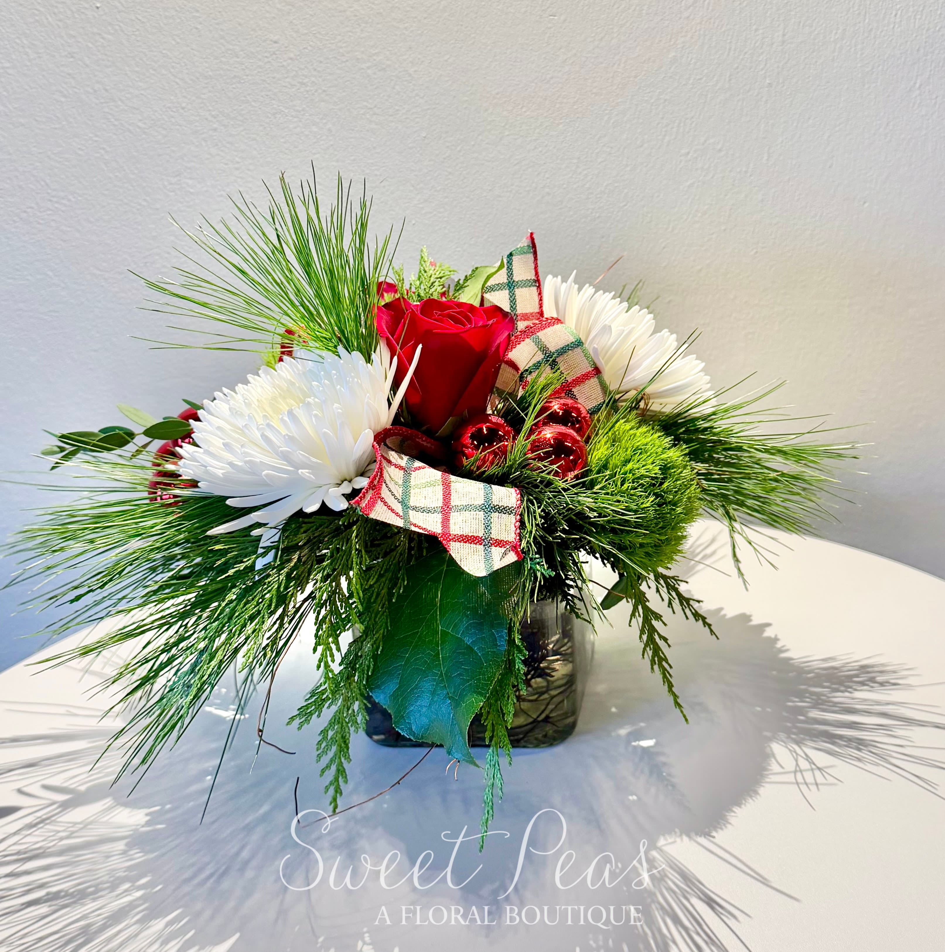 Holiday Cheer - Bring the warmth and charm of the season into any space with our Holiday Cheer arrangement. Designed in a modern glass vase, this festive piece features a lush bed of winter greens—including pine and cedar—creating a fresh, fragrant foundation.  At the heart of the arrangement sits red roses, beautifully complemented by crisp white spider mums and vibrant green trick dianthus. Accents of seasonal berries and a plaid holiday ribbon add playful texture and color, while small red jingle bells provide a whimsical finishing touch.  Perfect for gifting or brightening your own home, this arrangement captures the magic of the holidays in a stylish, compact design.