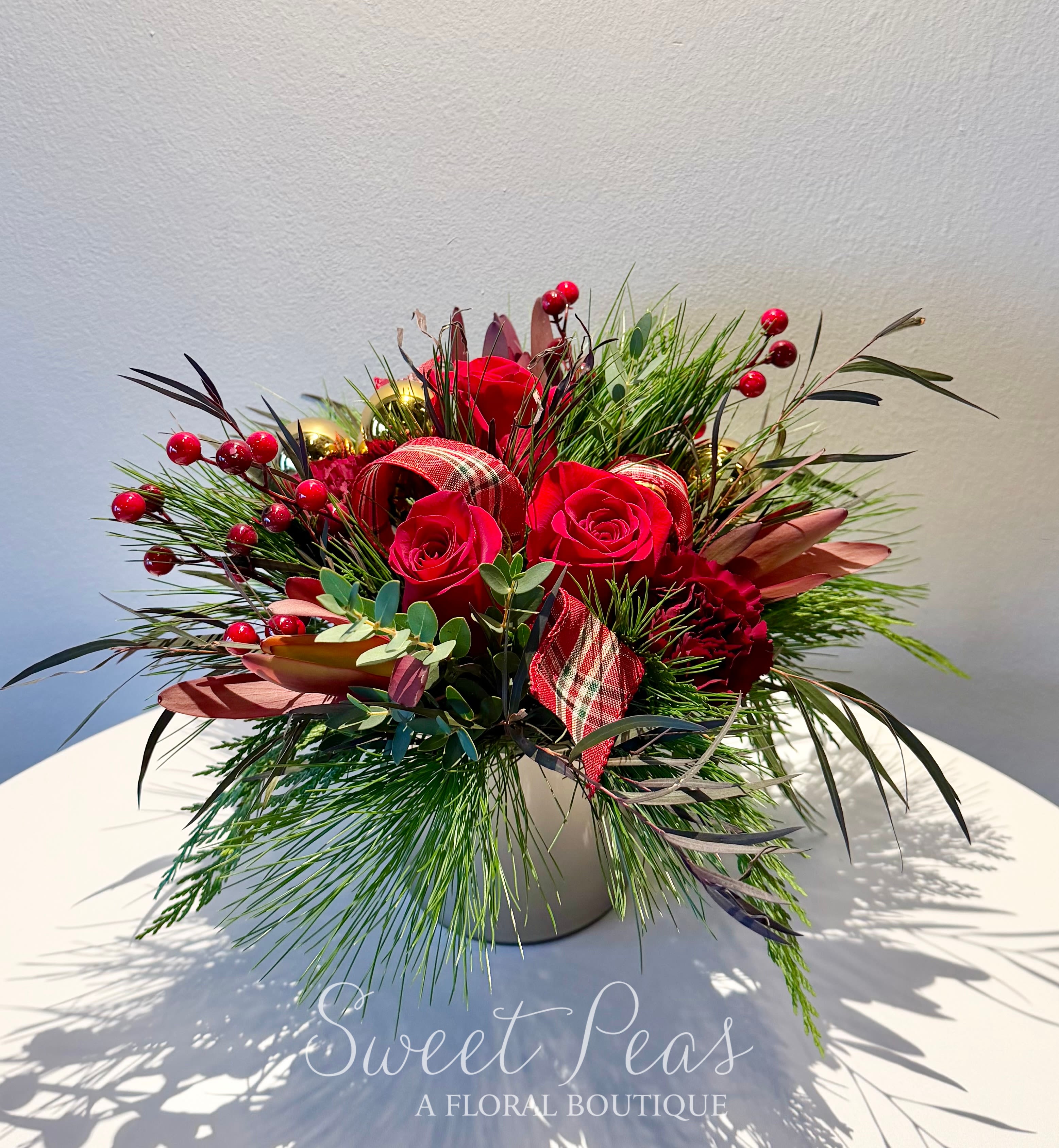 Crimson Noel - Our Crimson Noel Arrangement is a sophisticated celebration of holiday elegance. Designed in a modern ceramic container, this piece features luxurious red roses and deep red blooms nestled among an artful mix of winter greenery.  Textural accents—such as glossy holiday berries, gold ornament touches, and rich foliage in burgundy and bronze tones—add depth and warmth. Loops of festive plaid ribbon weave throughout, bringing a classic seasonal charm to the lush, airy composition.  This arrangement strikes the perfect balance between timeless holiday tradition and contemporary style, making it an ideal gift, centerpiece, or festive accent for any home or office.