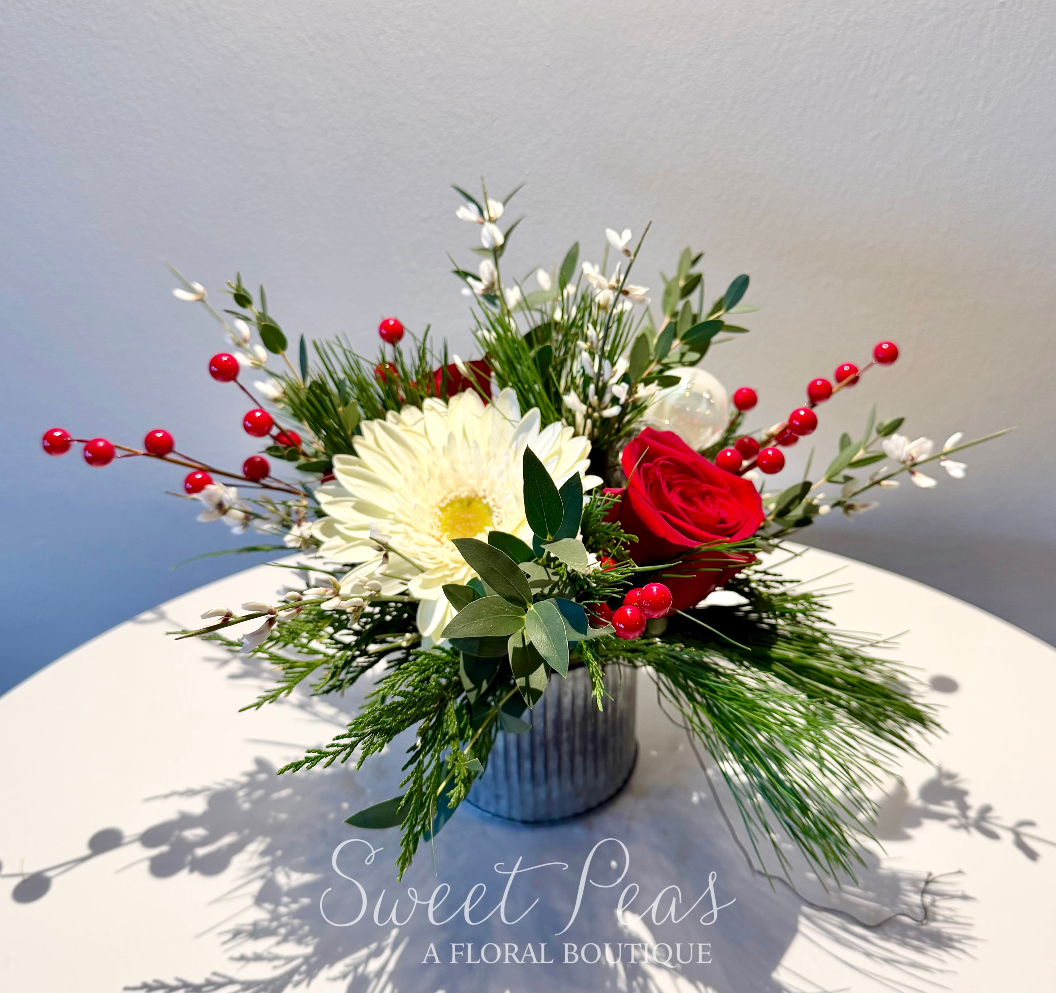 Winter Wishes - Bright, fresh, and full of seasonal cheer, our Winter Wishes Bouquet brings a modern touch to classic holiday florals. Designed in a galvanized metal container, this arrangement features a beautiful pairing of red roses and a crisp white gerbera daisy as the focal blooms.  Festive accents of red winter berries, soft white waxflower, and lush mixed evergreens create a natural, airy feel. Touches of eucalyptus add elegance, while a cluster of white and clear ornaments nestled among the greens brings a subtle hint of holiday sparkle.  Perfect for gifting, decorating, or sending warm wishes, this arrangement captures the joy and simplicity of the winter season.