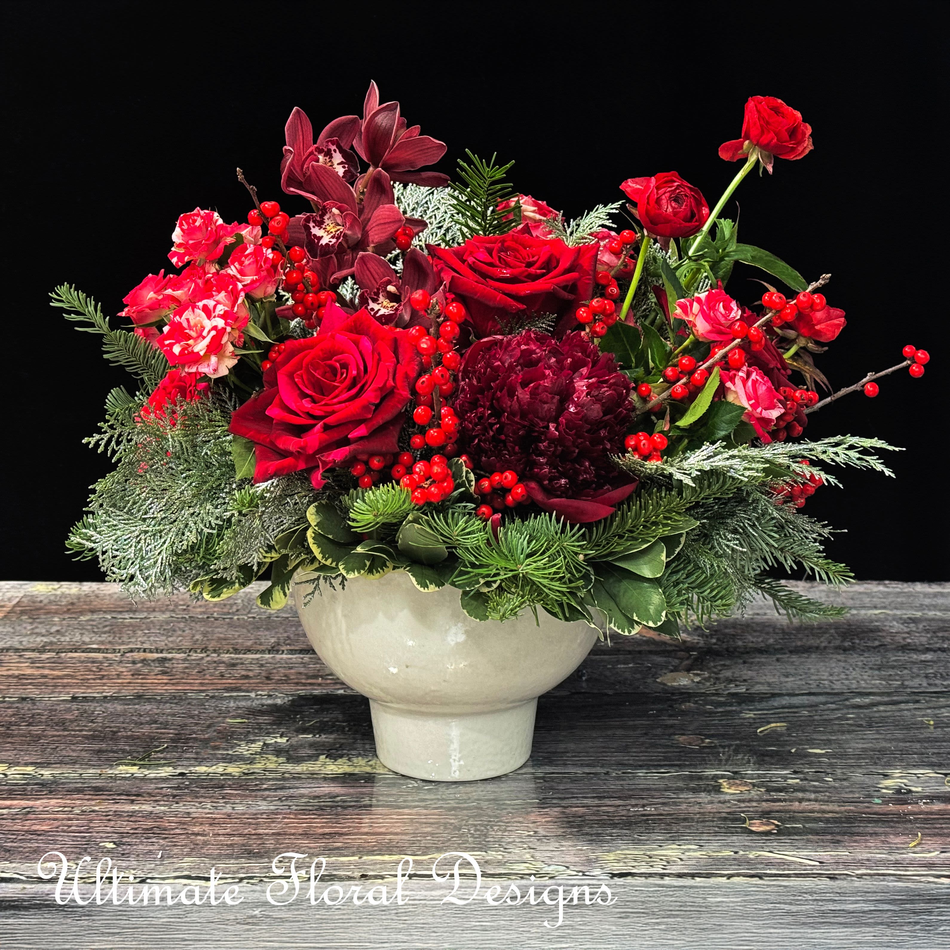  Crimson Winter Delight - This striking floral arrangement captures the festive elegance of the holiday season. Featuring a vibrant combination of crimson roses, dark cymbidium orchids, deep red ranunculus, and lush peonies, it evokes warmth and celebration. The seasonal touch of evergreen sprigs and frosted foliage adds a hint of winter charm, while bright red hypericum berries bring a festive sparkle to the design. Arranged in a timeless white ceramic vase, this piece is perfect for holiday gatherings, special occasions, or as a luxurious centerpiece to enhance your winter décor.