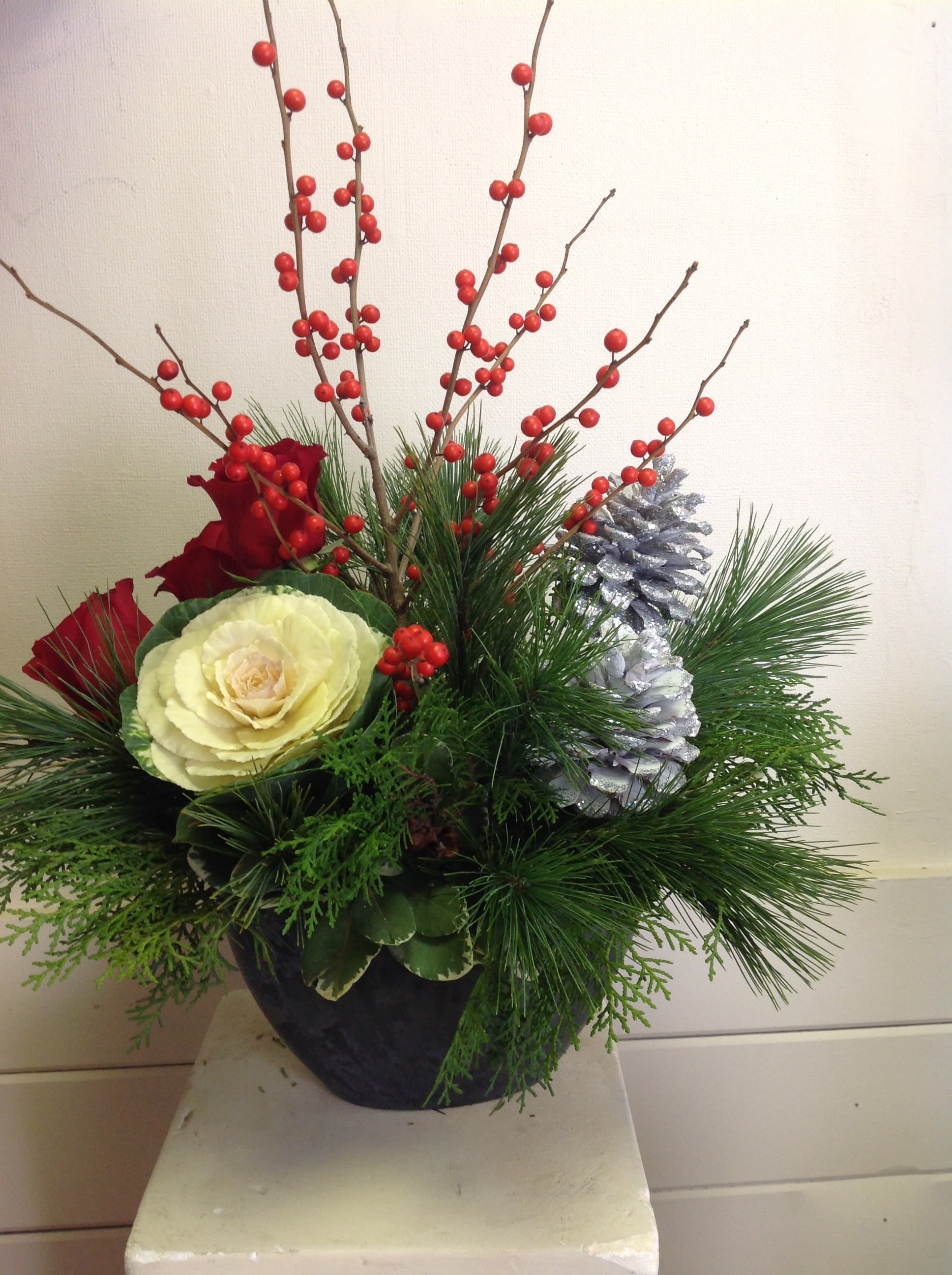 Holiday Cheer - Sleek modern container filled with red ilex berries, red roses, ornamental kale, mixed pines and silver tipped pine cones.