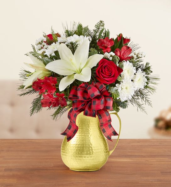 Country Christmas -  Arrangement with red roses and alstroemeria white Asiatic lilies, cushion poms and statice; accented with assorted Christmas greenery Artistically designed in our Hammered Bell Pitcher with burnished mirror-polished finish and a distinctive handle; pairs beautifully with any tableware; the perfect accent piece for both formal and