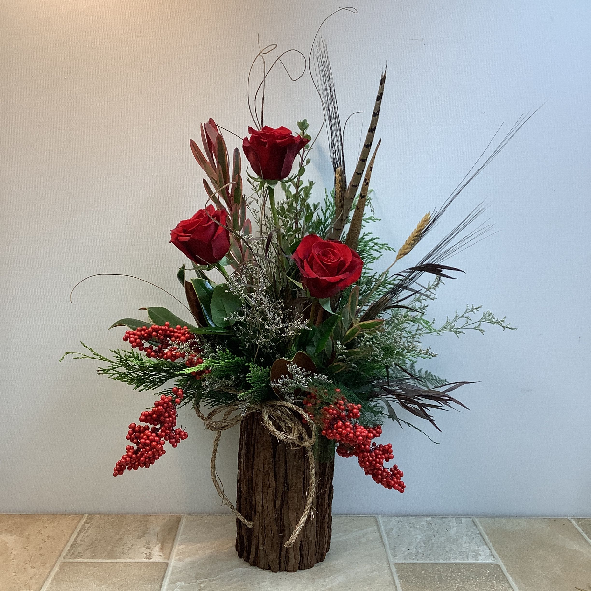 Woodland Serenade - This tall floral arrangement is presented in a bark-covered, cylindrical vase tied with twine. Gorgeous red roses, red silk berries and dramatic leucadendron offer vibrant color; while peasant feathers, wheat stems, curly brown ting ting branches, and lush green foliage provide an earthy balance. Rustic, yet elegant, this arrangement can be ideal for any occasion. Approximate size 17" w x 27" h.  Primary elements: rose, berry, feather  Flower delivery La Crosse, Onalaska, Holmen, West Salem, La Crescent. Flower shop. Plant store. Best florist La Crosse Wisconsin area.  Seasonal and holiday floral creations. Funeral arrangements. New baby bouquets. Birthday celebration flowers. Get well arrangements. Just because gifts. 