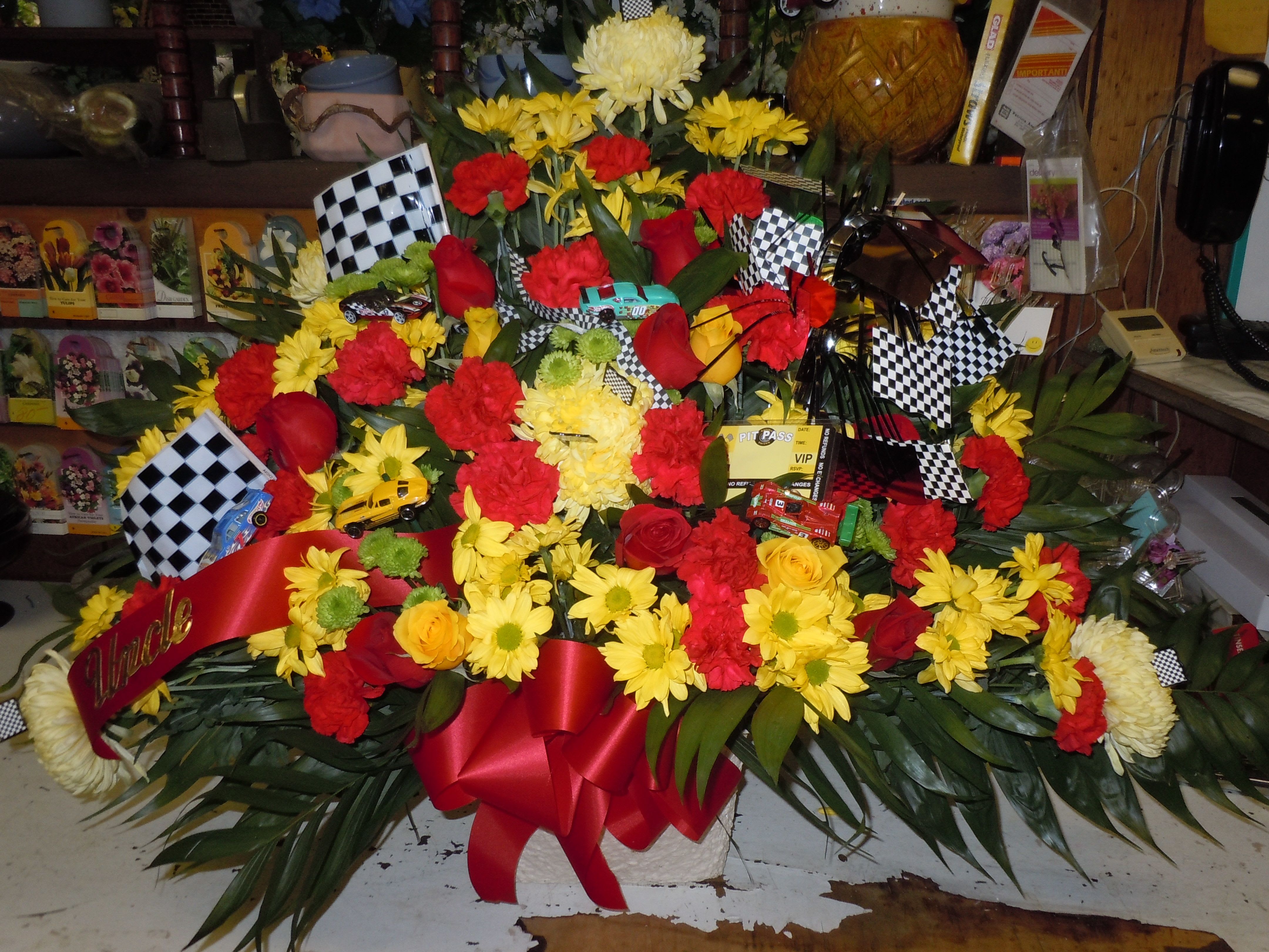 TB24 Sympathy Basket - A mixture of yellow daisies, red carnations, red roses, yellow fuji mums, baby's breath, green button poms, greens and red bow  **Flags and race cars supplied by customer**  Banner - $15.00 + extra  Substitutions may apply due to flower availability