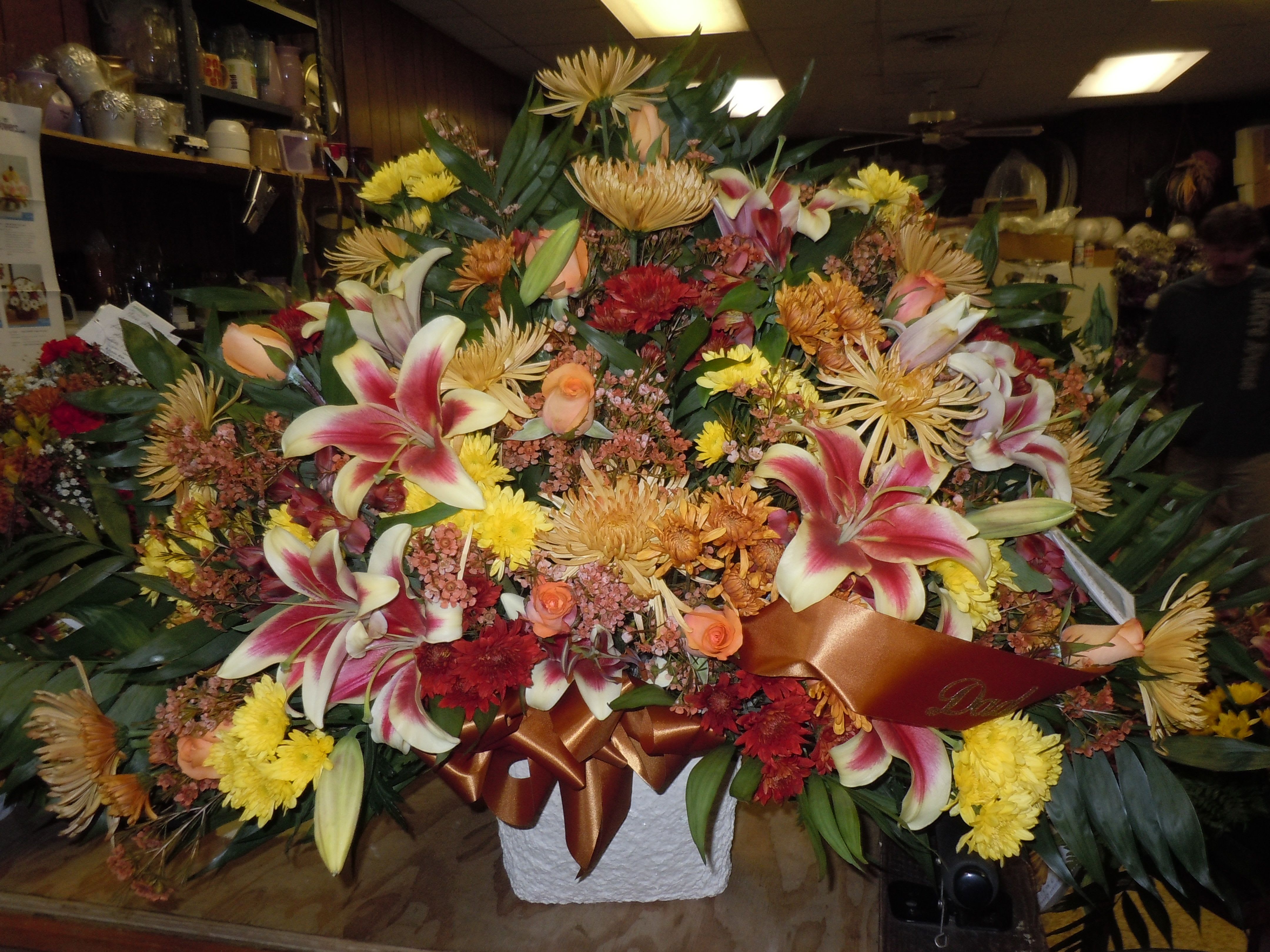 TB25 Sympathy Basket - A mixture of fall bronze and yellow cushion, and burgandy daisies, orange roses, bronze fuji mums, shocking lilies, red alstromaria, orange wax flower and bronze bow  Banner - $15.00 + extra  Substitutions may apply due to flower availability