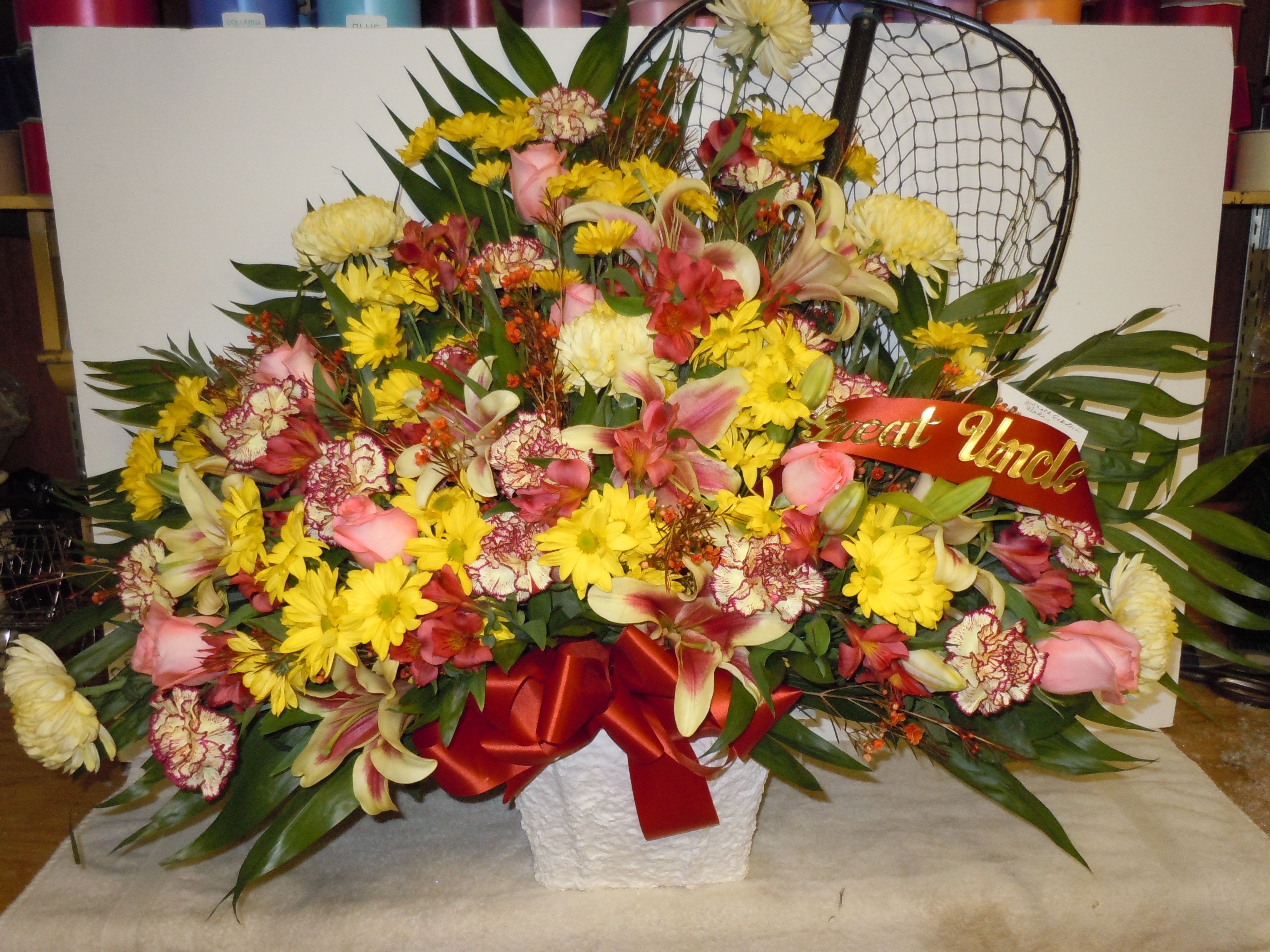 TB26 Sympathy Basket - A mixture of yellow daisies, yellow carnations tipped burgandy, orange roses,  yellow fuji mums, red wax flower, red alstromaria, shocking liles, greens and red bow   Fishing net supplied by customer  Banner - $15.00 + extra  Substitutions may apply due to flower availability