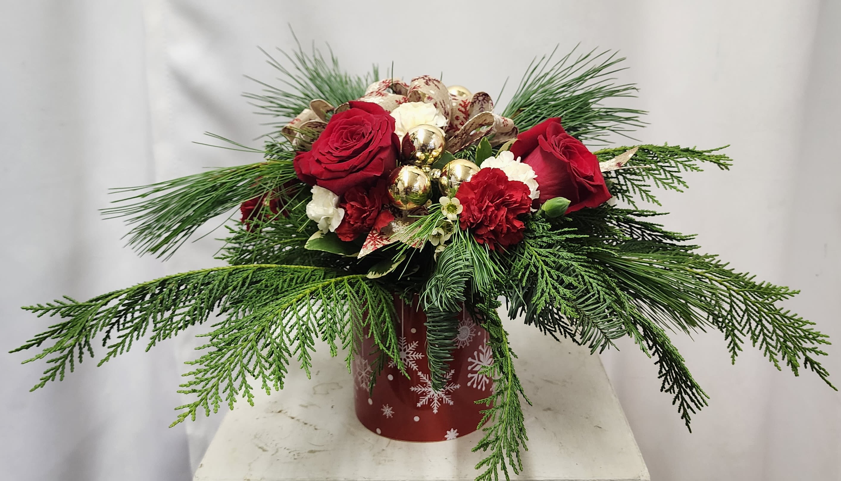 Snowflake Charm - Celebrate the magic of the holidays with Snowflake Charm—a simple and elegant display featuring red roses, red and white mini carnations, elegant magnolia leaves, and lush holiday greens. Sparkling gold ornaments and a coordinating holiday bow add a touch of glamour, making this arrangement the perfect centerpiece for seasonal gatherings or a thoughtful gift to brighten any winter day.