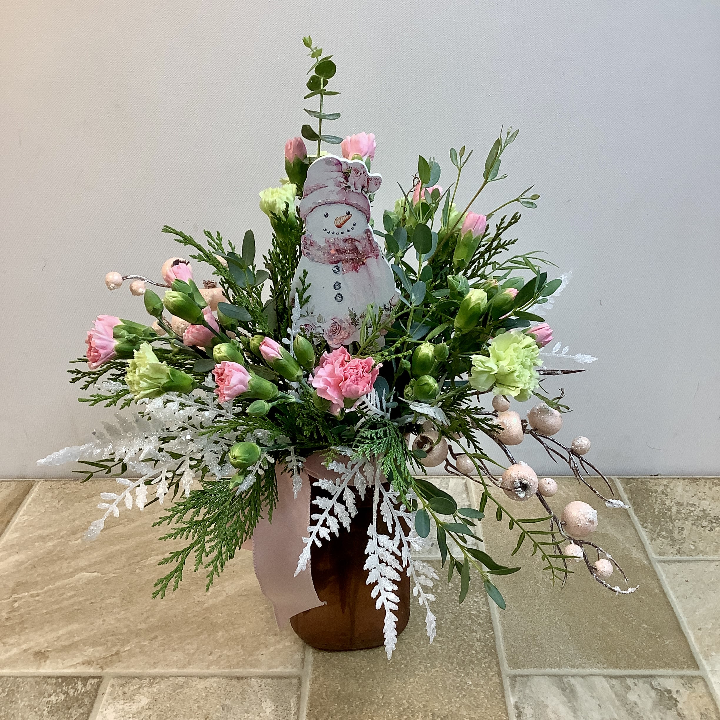 Frosty's Frolic - This cheerful winter arrangement featuring a keepsake snowman ornament is sure to bring a smile. Surrounding the snowman are delicate pink and green mini carnations, accented with sprigs of cedar, eucalyptus, and frosted white fern. Glittery pink berries add a hint of sparkle, while the rosy-colored vase and soft pink ribbon bring warmth and sophistication to the wintry theme. Approximate size: 13" w x 15" h  Primary flowers: carnation  Flower delivery La Crosse, Onalaska, Holmen, West Salem, La Crescent. Flower shop. Plant store. Best florist La Crosse Wisconsin area.  Seasonal and holiday floral creations. Funeral arrangements. New baby bouquets. Birthday celebration flowers. Get well arrangements. Just because gifts.