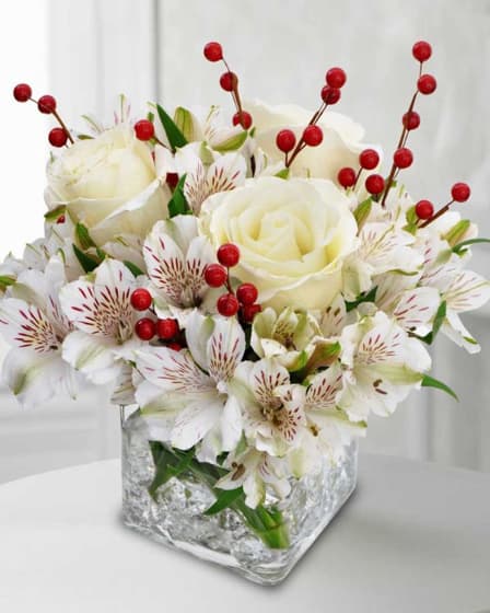 Rudolph the Reindeer - Rudolph the Reindeer features white alstroemeria, crisp white roses, and red berry strands symbolizing Rudolph’s bright red nose in the night sky arranged in a clear cube vase.  