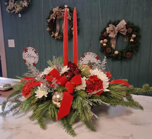 Double Candle Christmas Centerpiece - Light up the season with our Double Candle Christmas Centerpiece. Two glowing candles create a warm, festive ambiance as they rise above a lush bed of fresh winter greens and long-lasting seasonal Christmas flowers. Accented with pinecones, rich holiday ribbons, and touches of classic Christmas charm, this centerpiece is perfect for dining tables, holiday gatherings, or sending warm wishes to someone special. A timeless and elegant way to celebrate the beauty of the season.