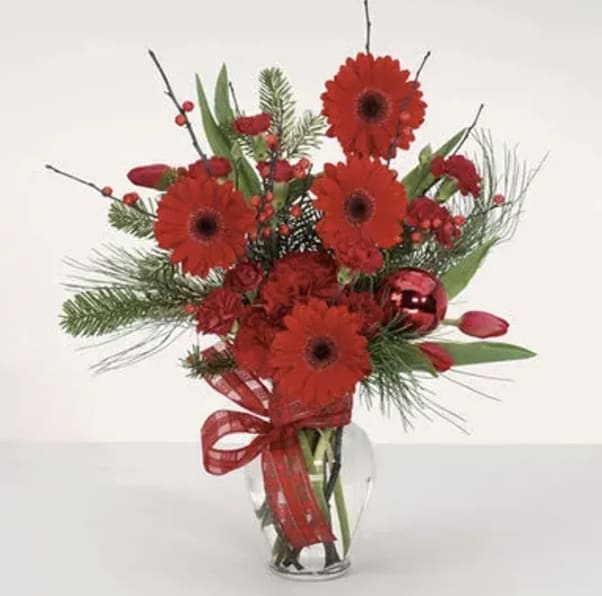 Color Me Christmas - Vibrant and full of holiday cheer, Color Me Christmas is a festive arrangement bursting with rich red tones and seasonal charm. Featuring bold red gerbera daisies, accented with winter greenery, red berry details, and a touch of holiday sparkle, this design instantly brings warmth and joy to any space.  Finished with a cheerful red ribbon and arranged in a clear glass vase, this bouquet is perfect for Christmas celebrations, hostess gifts, or adding a bright seasonal touch to your home or office. Color Me Christmas is a joyful expression of the season—classic, colorful, and unmistakably festive.
