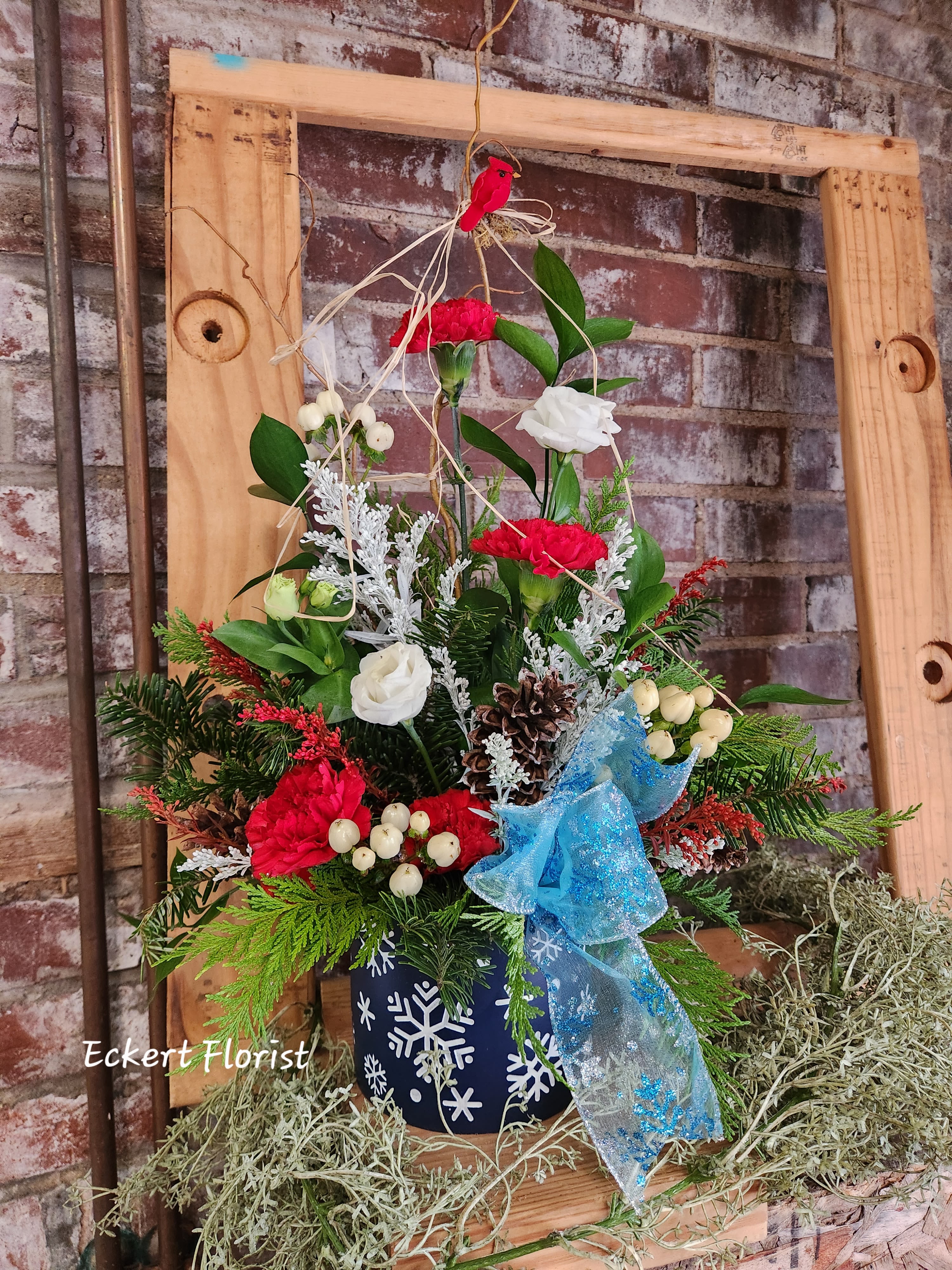 Eckert Florist's Festive Snowflake Arrangement *Our Local Delivery Only - Designed in a blue ceramic snowflake container, this arrangement is filled with red and white fresh blooms with a mix of pine cones and winter greens. Designer ribbon is added to complete this winter look. Container measures approx. 4" Diameter x 4" High *Our local delivery only
