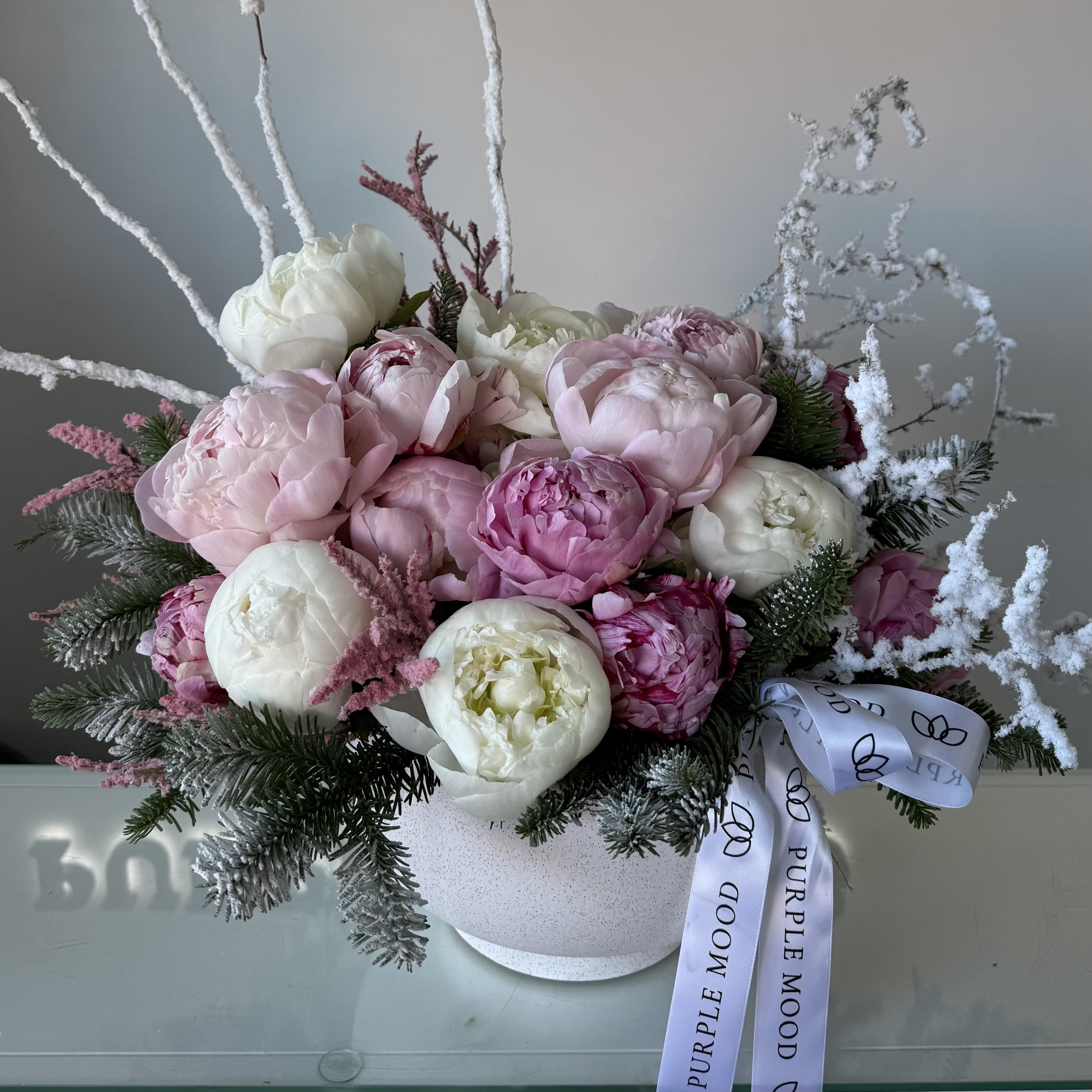 Snowy Peony vase - Indulge in winter elegance with our Snowy Peony Vase, a luxurious arrangement designed to capture the softness of freshly fallen snow. Featuring premium blush and white peonies, fluffy winter greens, and frosted branches, this piece blends romance with a serene seasonal charm.  Artfully arranged in a modern white vase and finished with our signature Purple Mood ribbon, it makes a stunning gift for the holidays, winter birthdays, or simply to bring a touch of magic into any space.  Perfect for: winter celebrations, thoughtful gifting, home décor, and special occasions. Vase included. 