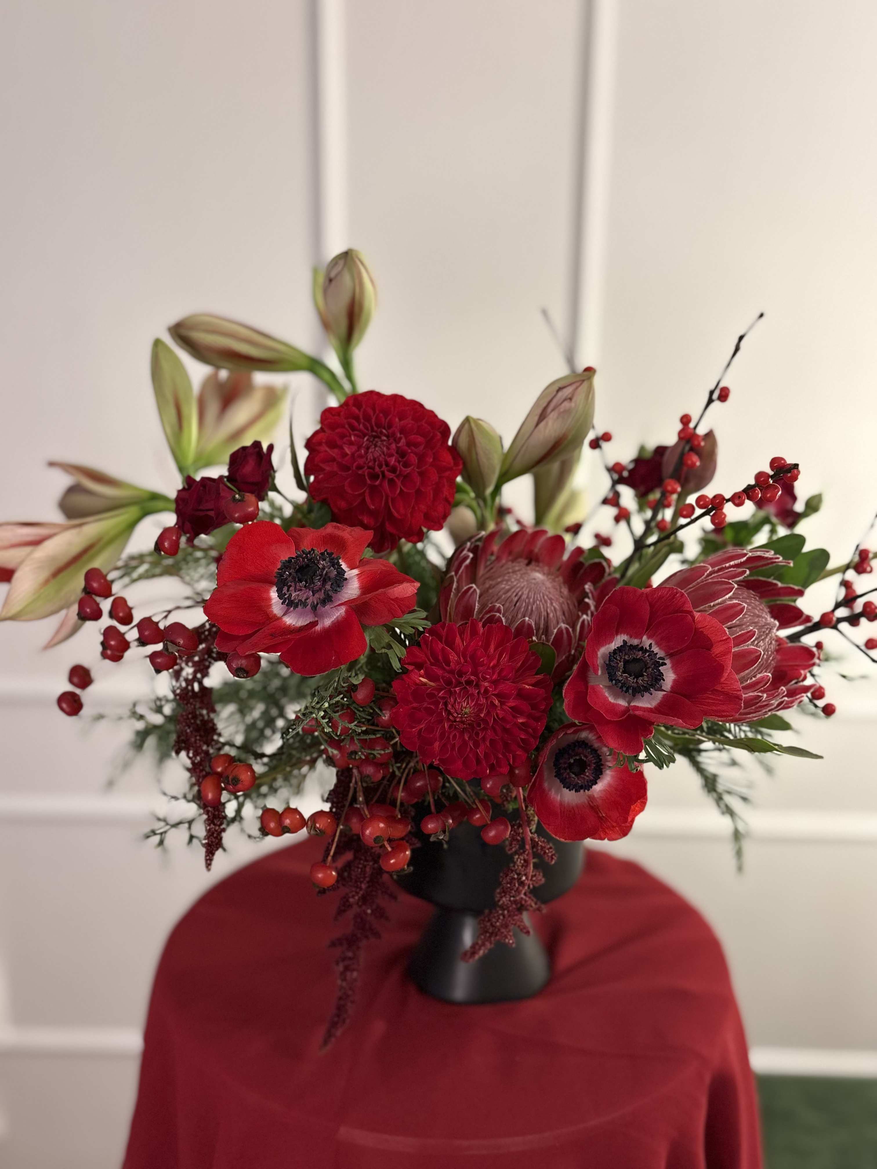 Sugar Plum Dreams - A rich red arrangements created with Christmas greens and a variety of red seasonal flowers, including amaryllis. Created in a black compote style vase.