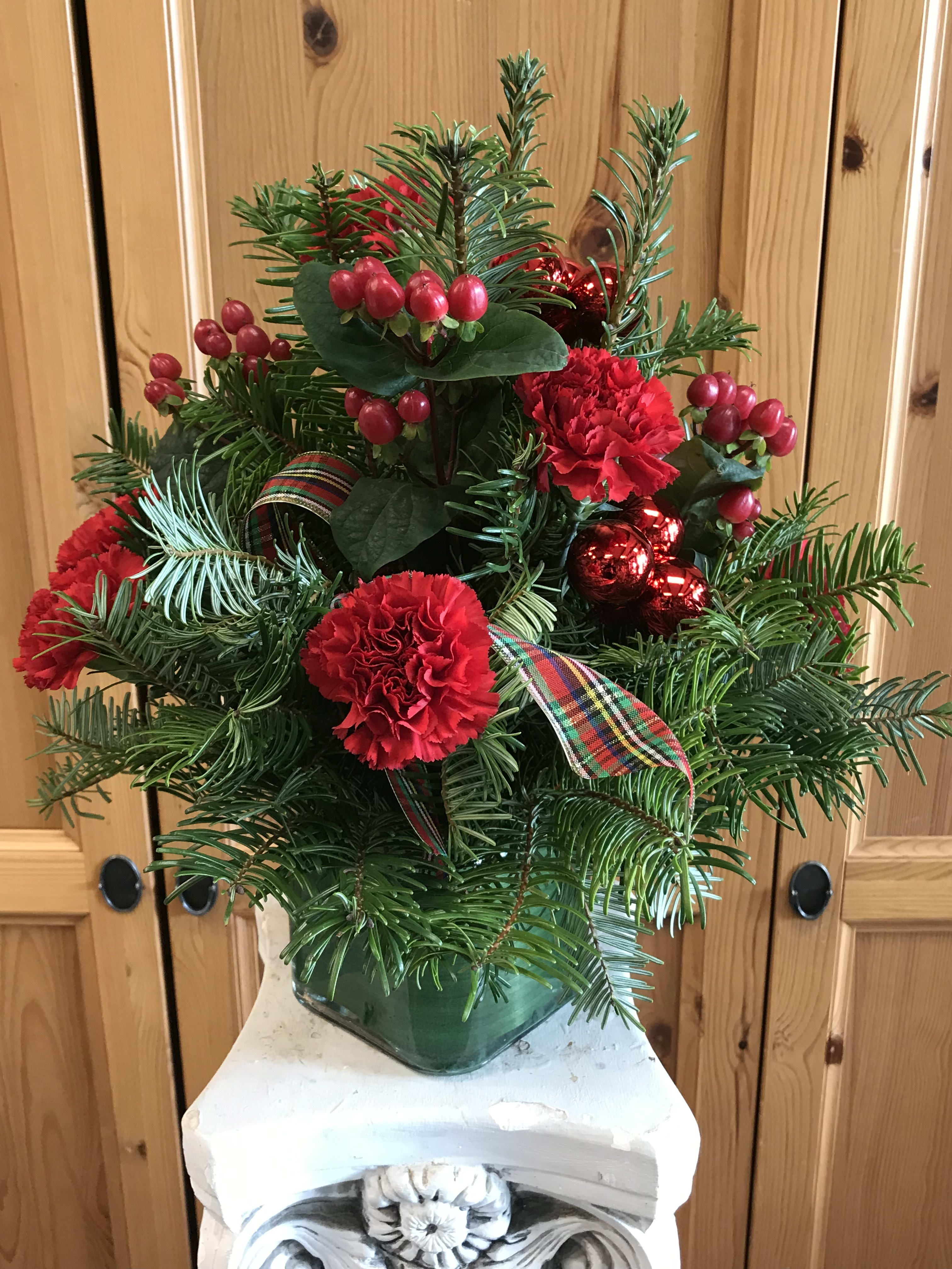 Holiday Cheer - A lush holiday arrangement of winter greens, red hypericum, red carnations, holiday ornaments, plaid ribbon. and pine cones.The perfect gift for yourself or your family.