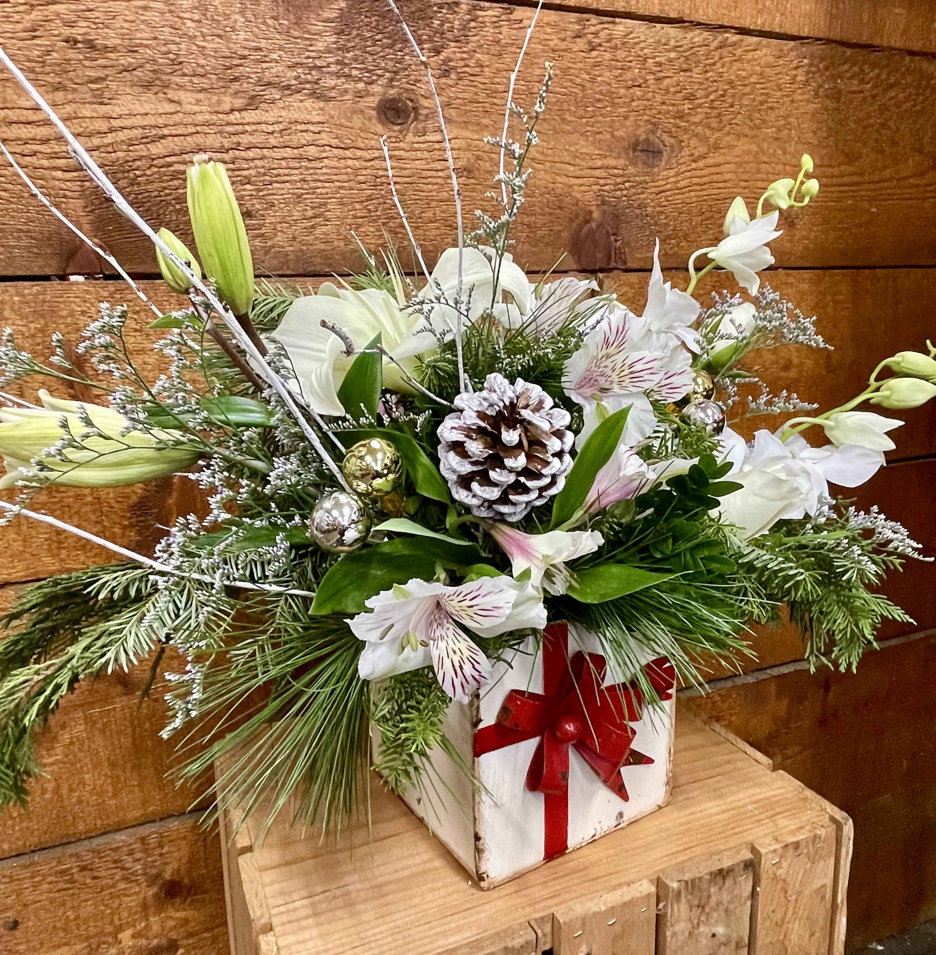 Gift of Giving - Send some holiday cheer with this seasonal arrangement. White lily, roses, alstroemeria, and orchids are designed in a vintage holiday tin box accompanied by Winter greens, pinecones, Ornaments and white sticks.