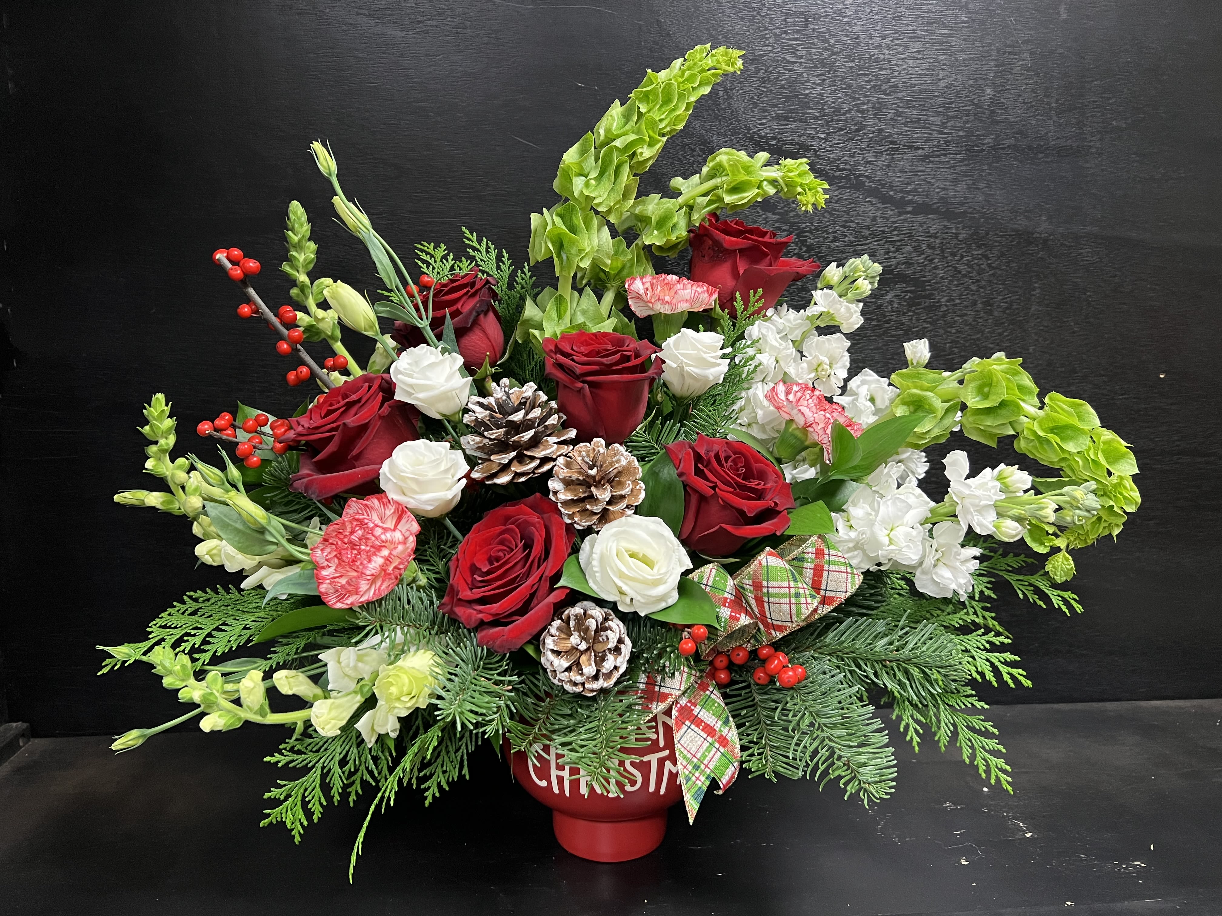 Merry Christmas  - This holiday flower arrangement is built in a Merry Christmas pot showcasing various Christmas greens, roses, snapdragons, stock, bells of Ireland, holly, pinecones,  carnations, and lisianthus.  