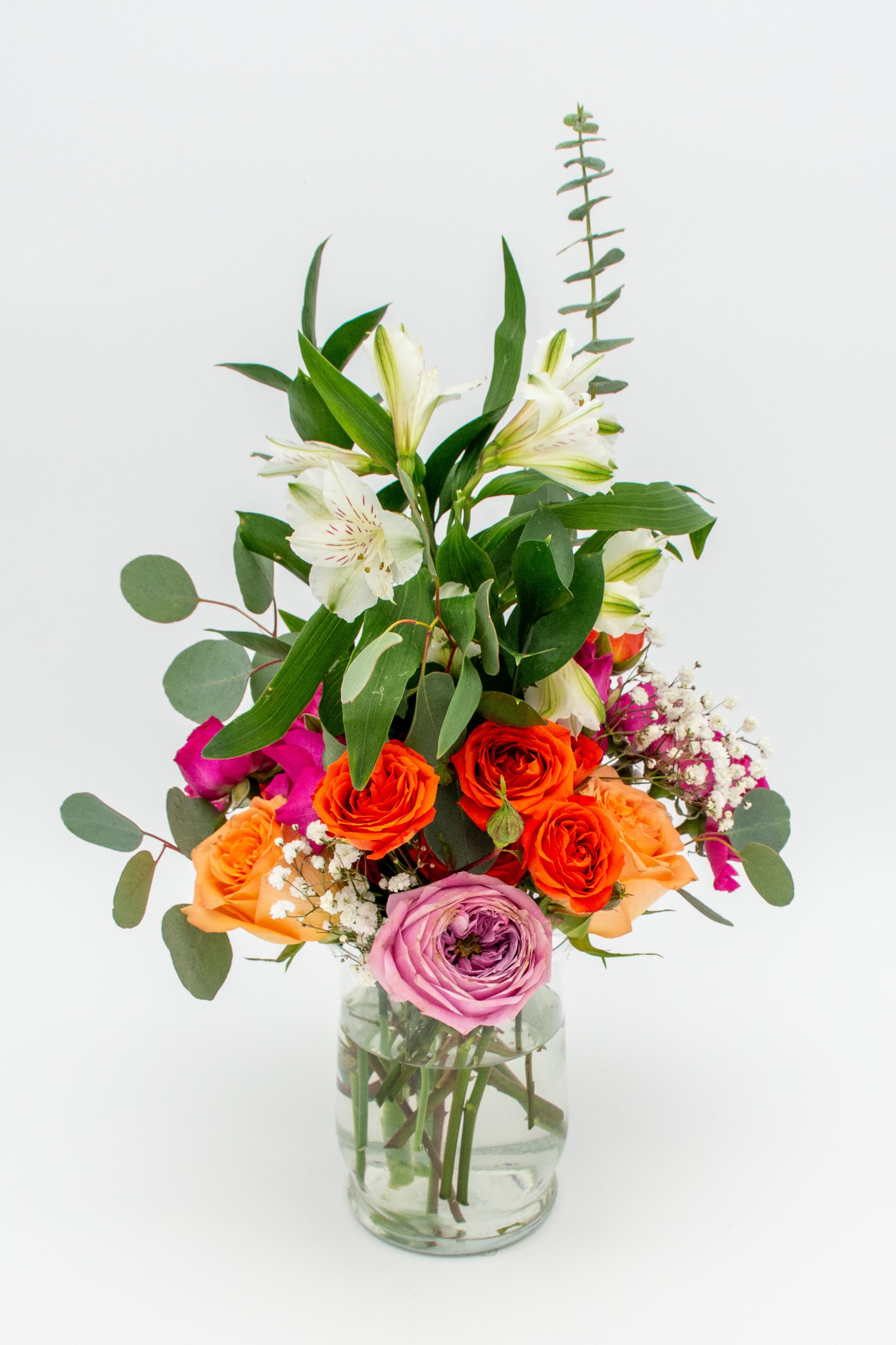  Sweet Sunset.     - A vertical and dynamic arrangement combining the energy of orange, fuchsia, and pink roses with the freshness of white alstroemerias and eucalyptus foliage, creating a clean, modern, and colorful design. Arranged in a transparent cylindrical vase that showcases the natural stems and adds a contemporary touch.