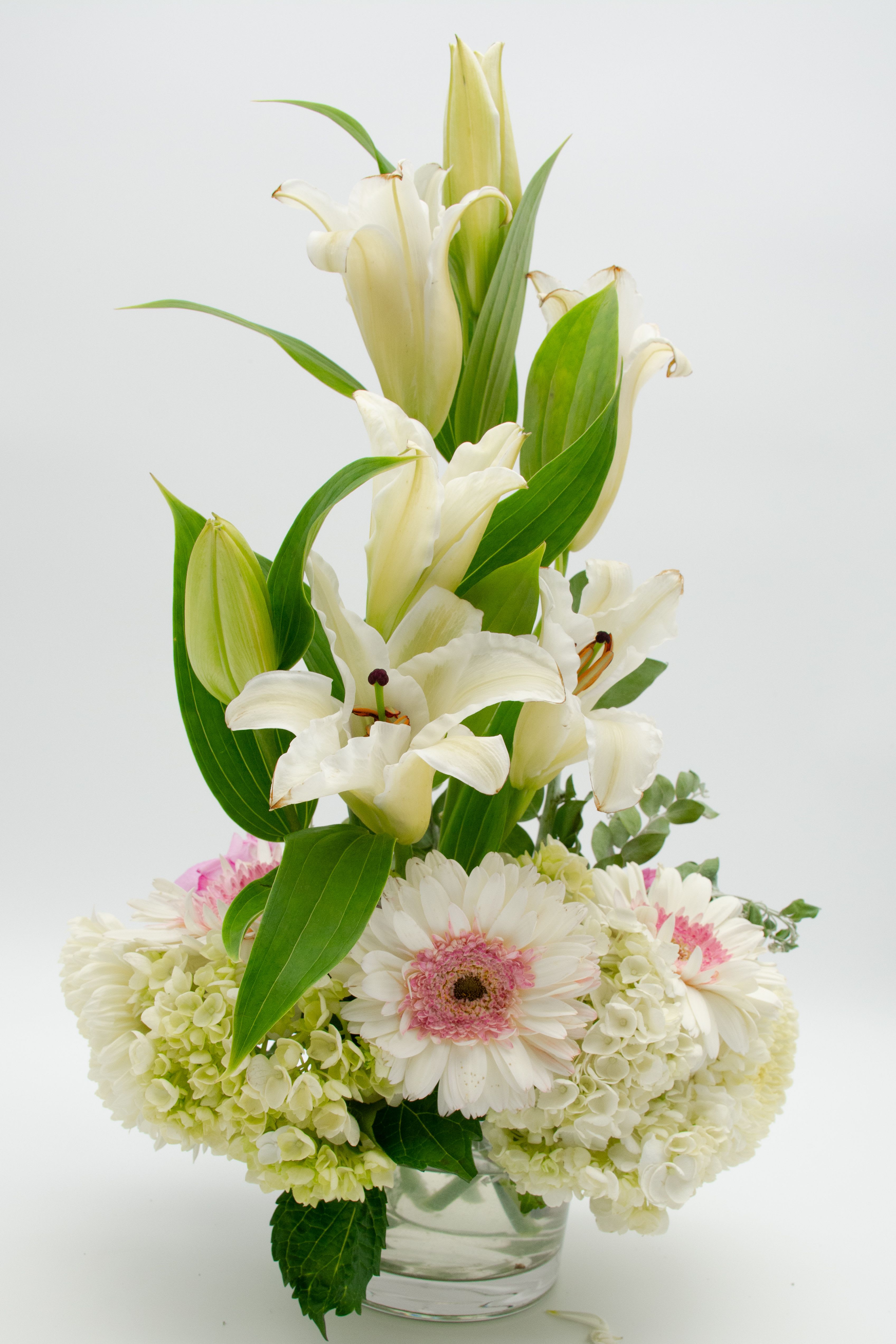  Snow Charm.  - A refined and luminous arrangement featuring elegant cream lilies in a vertical structure, paired with a lush base of white hydrangeas, soft-tone gerberas, and delicate carnations. The composition creates a serene, fresh, and balanced effect.