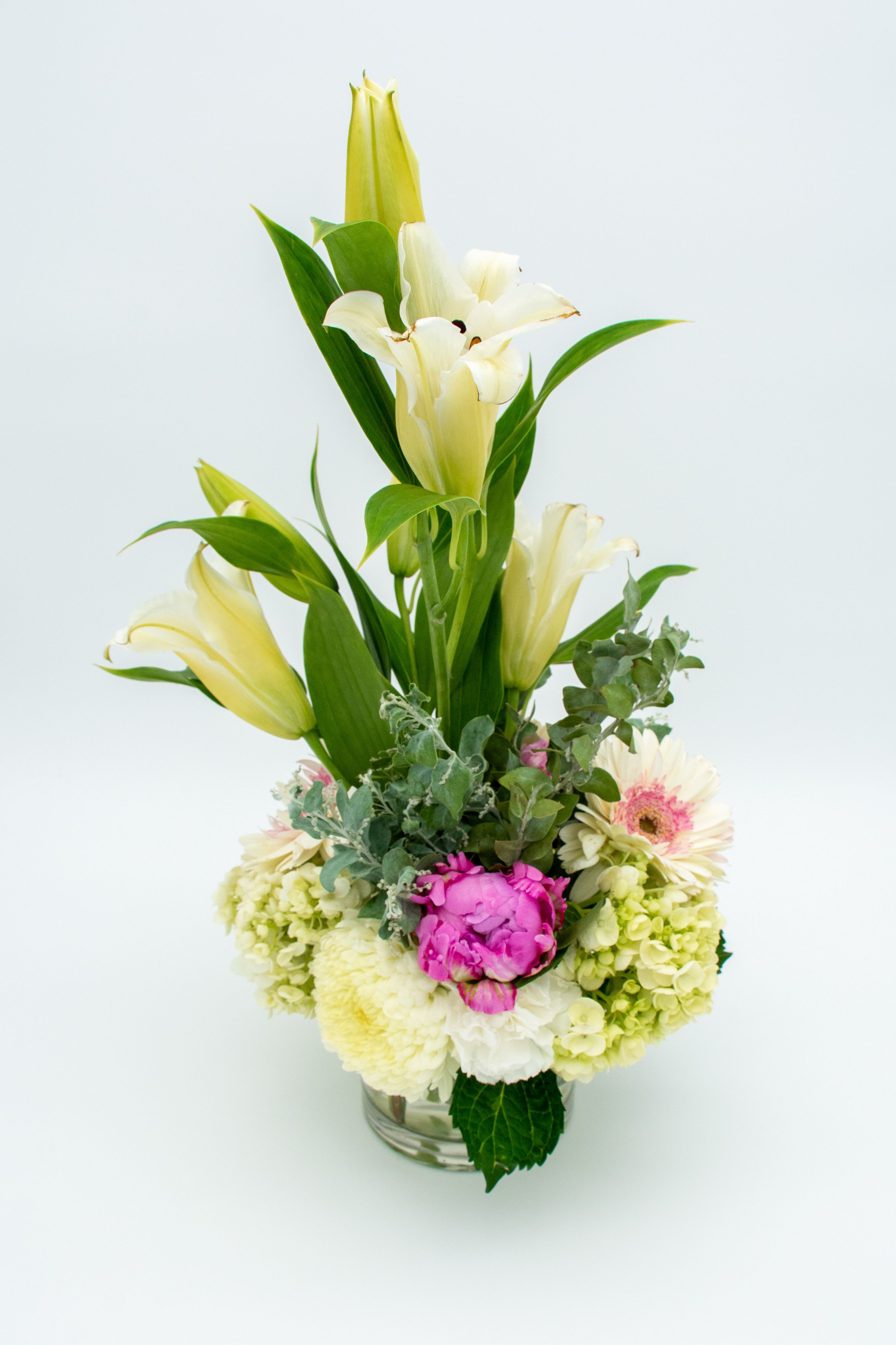 Spring Light  - A modern and elegant floral arrangement featuring white Oriental lilies rising in vertical lines, adding height and sophistication. At the base, a harmonious blend of cream hydrangeas, pale pink gerberas, light carnations or chrysanthemums, soft dusty-miller foliage, and a vibrant fuchsia peony that brings contrast and life to the design. Presented in a transparent vase that highlights the purity of the composition. A fresh, luminous design perfect for special occasions, modern interiors, or delicate celebrations.