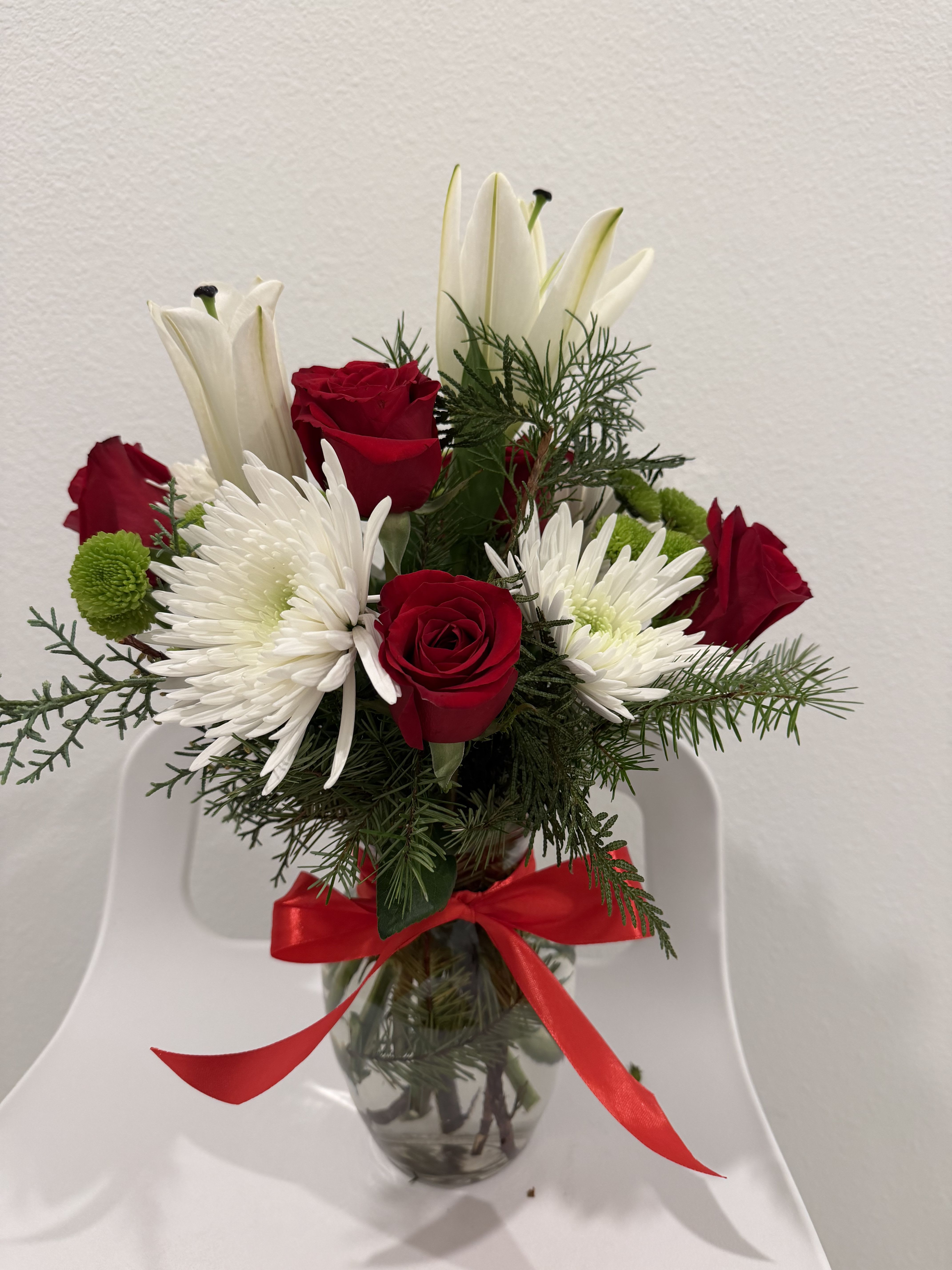 Holiday Glow Arrangement - A festive mix of red roses, white lilies, bright mums, and winter greens, tied with a radiant red bow. A cheerful holiday arrangement that brings a warm seasonal glow to any space.