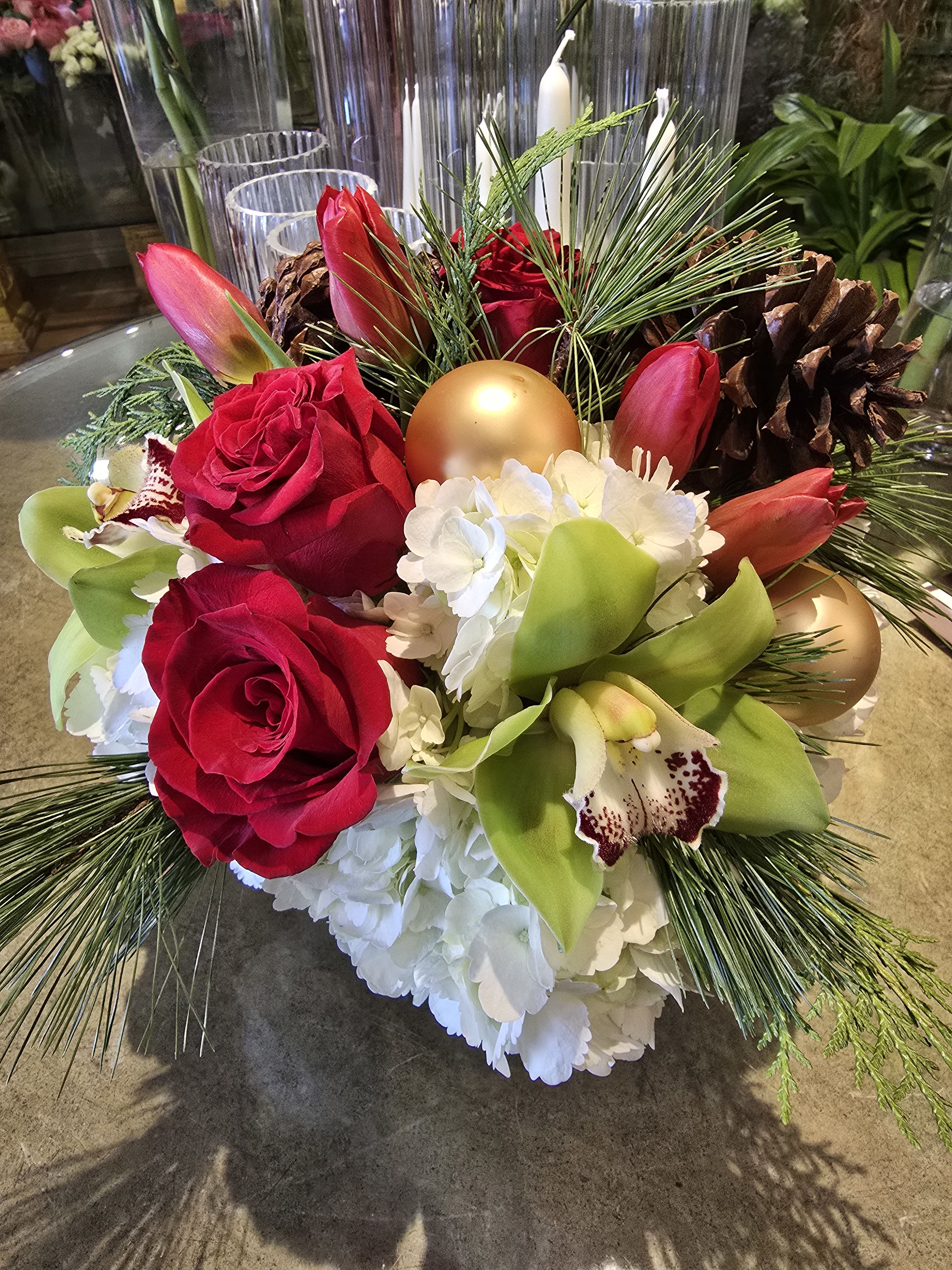 Festive Red &amp; Green Centerpiece - This elegant Christmas floral arrangement combines classic holiday colors with luxurious seasonal blooms. Featuring vibrant red roses, festive red tulips, lush white hydrangeas, and striking green cymbidium orchids, this arrangement is accented with natural pine branches, pinecones, and metallic gold ornaments for a rich, festive touch. Perfect as a holiday centerpiece, Christmas table decoration, or winter gift arrangement, this design brings warmth and sophistication to any space. Ideal for those searching for luxury Christmas flowers, holiday floral décor, or premium winter flower arrangements, this piece captures the magic and elegance of the season.