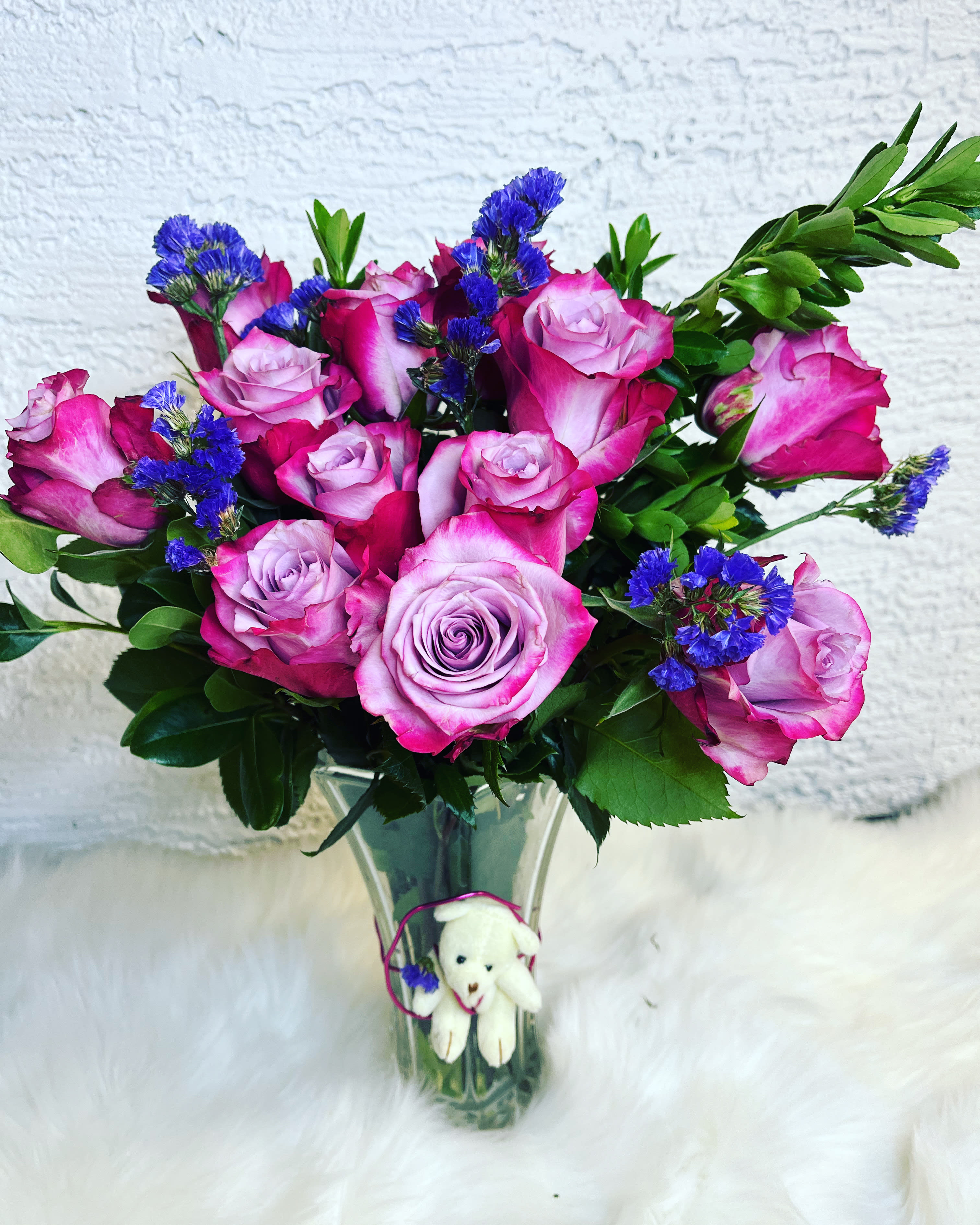  Affectionate - A vibrant and romantic arrangement featuring 12 stunning purple roses, accented with pops of fresh purple statice and lush greenery. Artfully arranged in a clear glass vase, this design is perfect for birthdays, anniversaries, or simply to brighten someone’s day. Includes a sweet plush bear charm for an extra touch of love.  Approximately 15” H x 12” W Flower types and accent colors may vary slightly based on seasonal availability. 